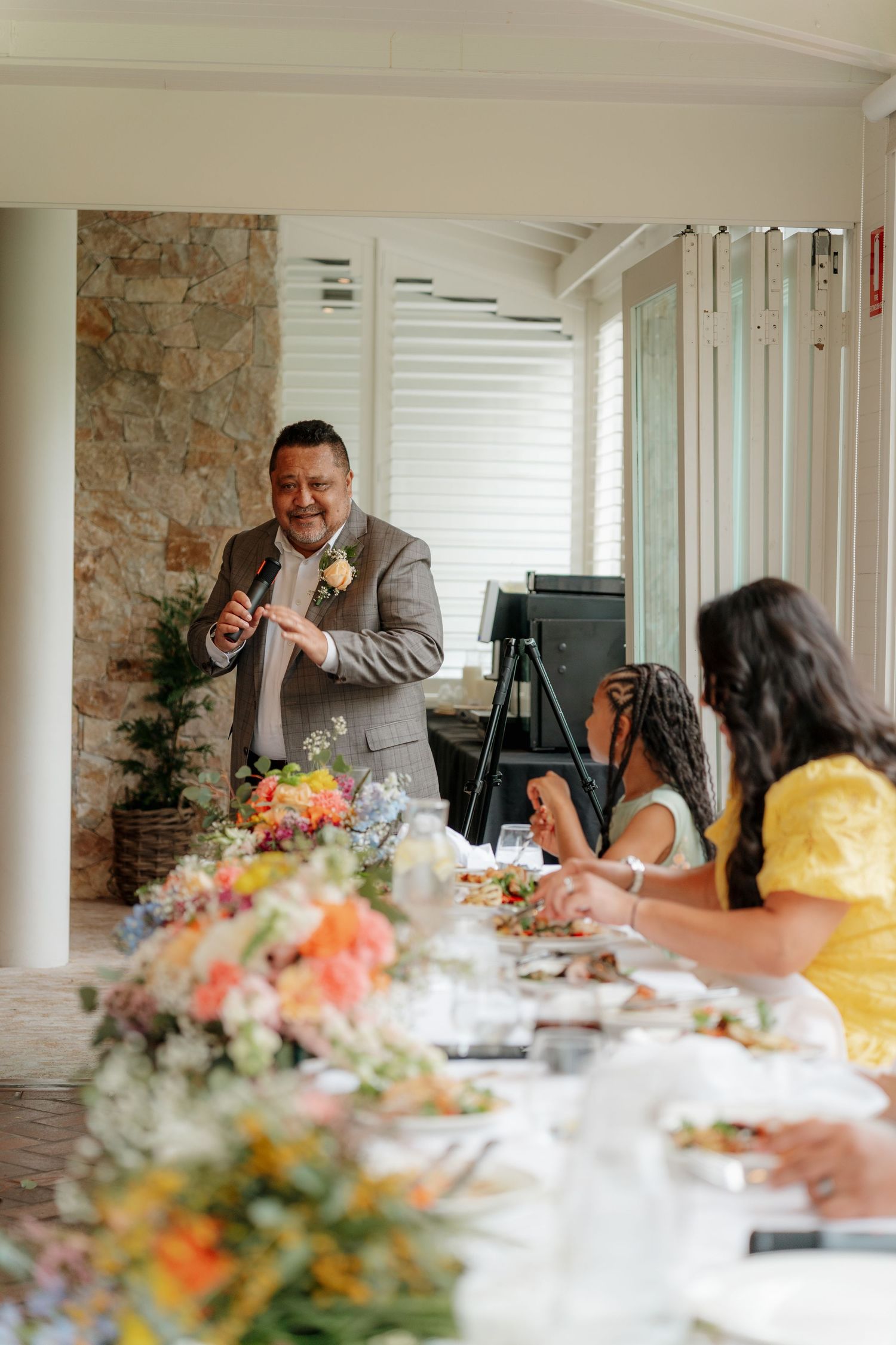 Man in brown suit giving a wedding speech at an elegant floral reception table with guests in yellow dresses.