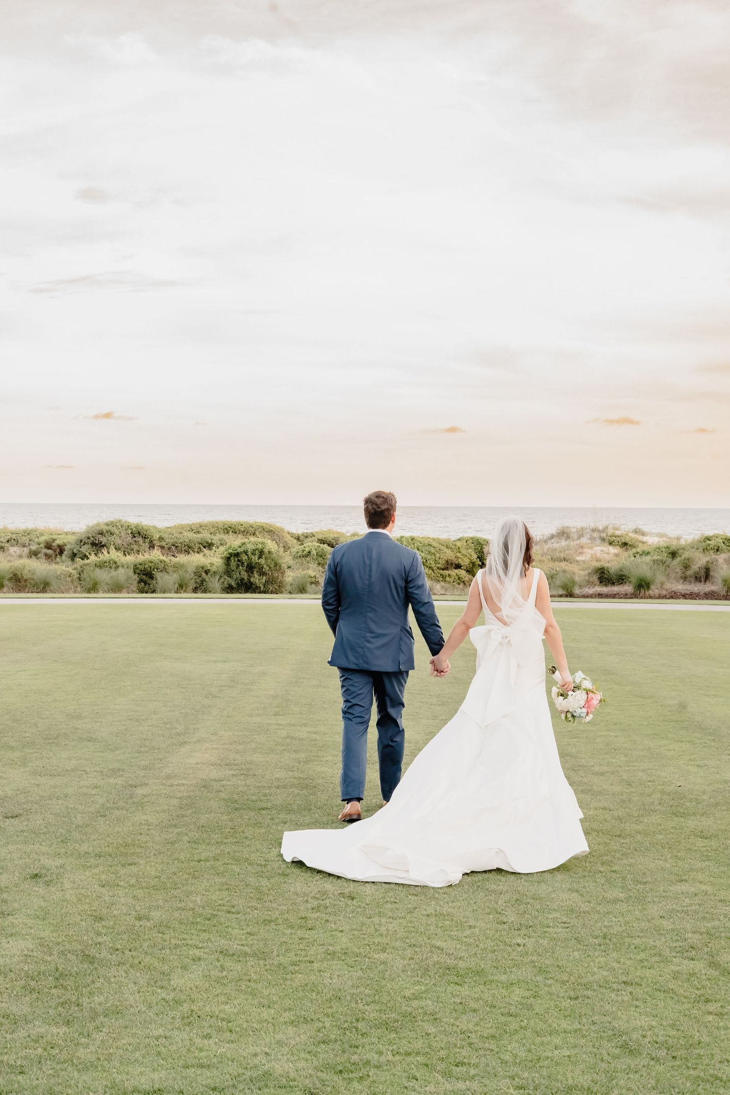 Bride and groom holding hands walking toward ocean on green lawn, bride in white gown with veil, groom in navy suit.