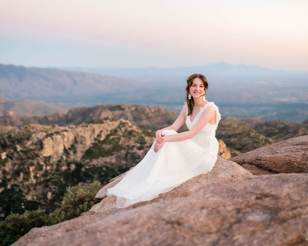 Bride in white gown sitting on rocky cliff with mountain valley view at sunset.