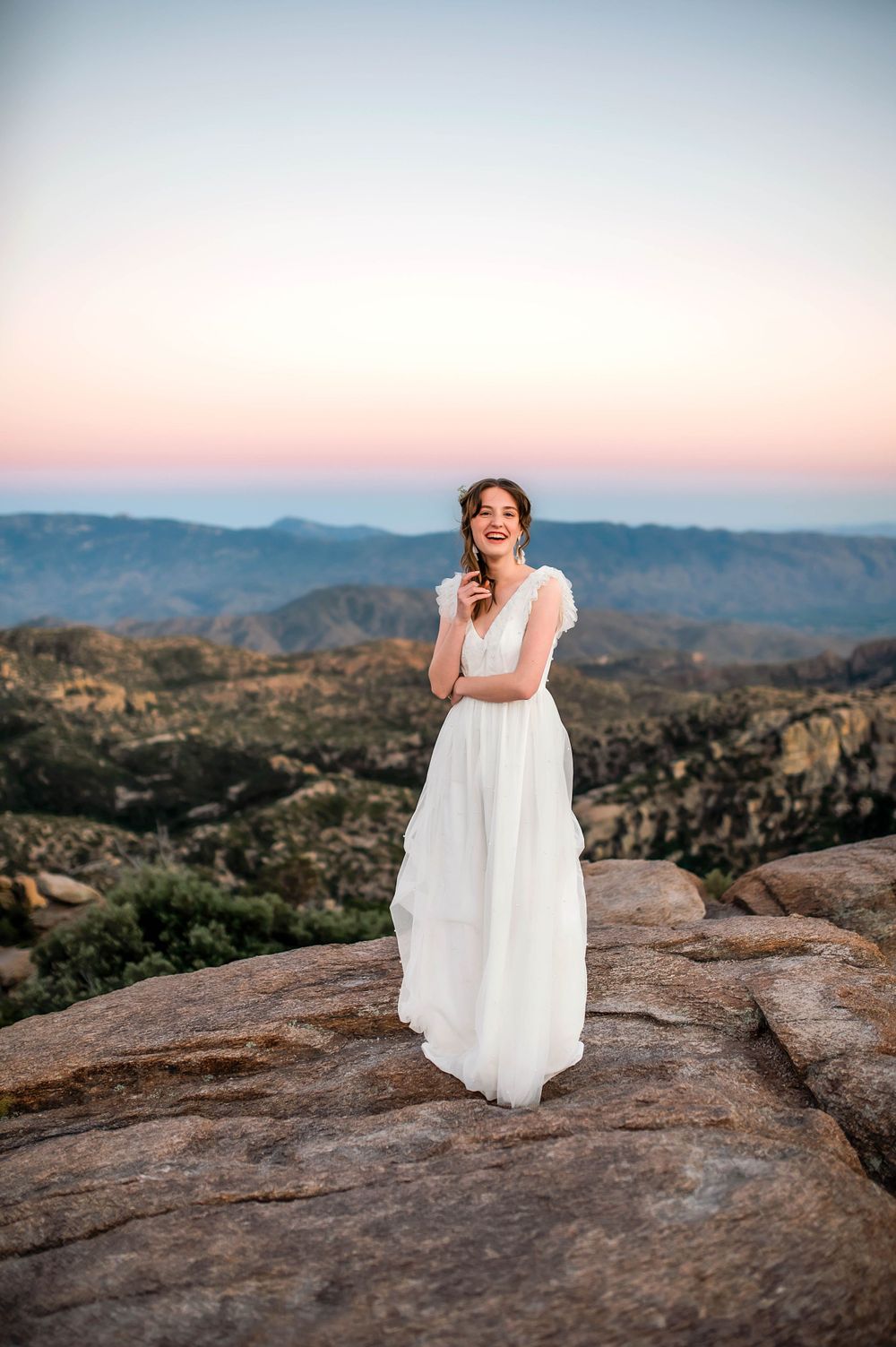 Bride in white gown standing on rocky mountain summit at sunset with panoramic valley views.