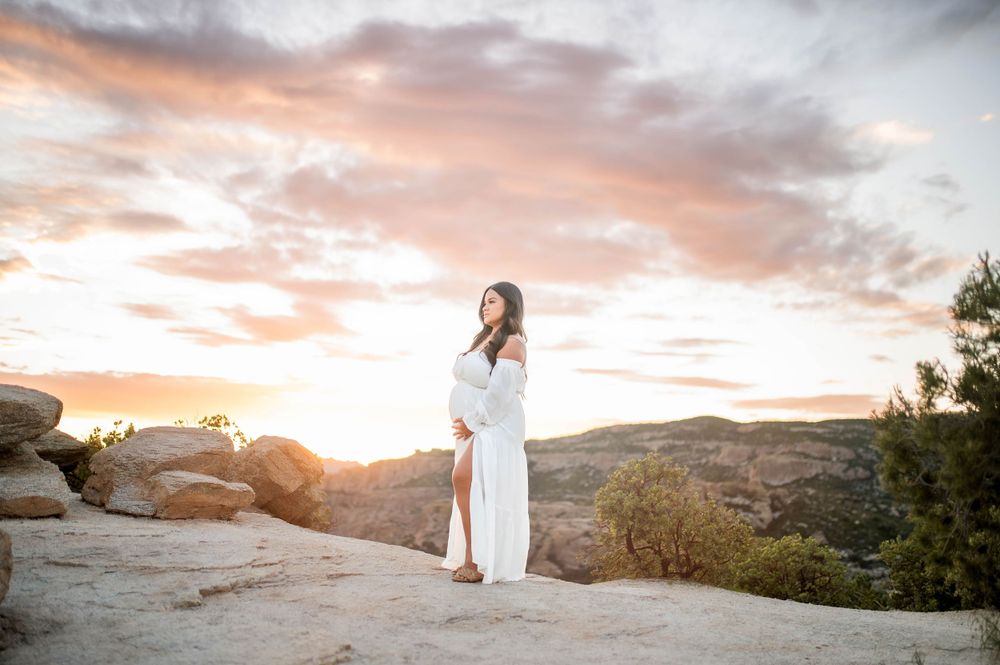 Pregnant woman in white off-shoulder gown posing on rocky terrain at sunset.
