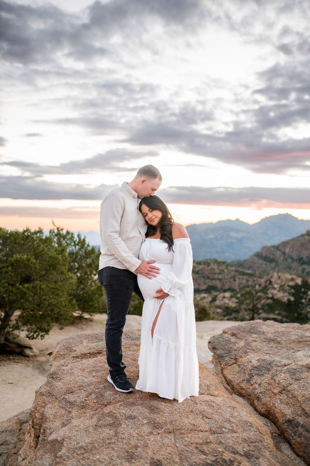 Couple embracing on rocky overlook at sunset, woman wearing white maternity gown.