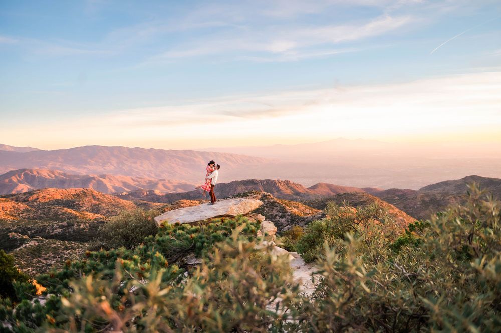Couple standing on mountain peak overlooking scenic valley landscape at sunset.