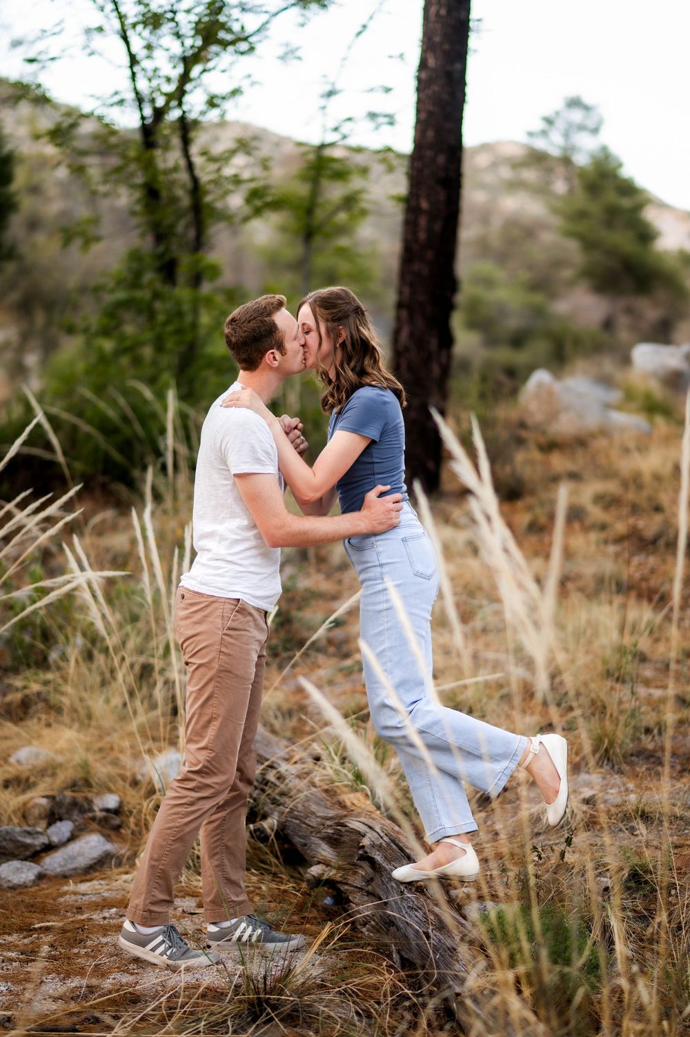 Couple shares a tender kiss while standing on a log in a scenic forest.