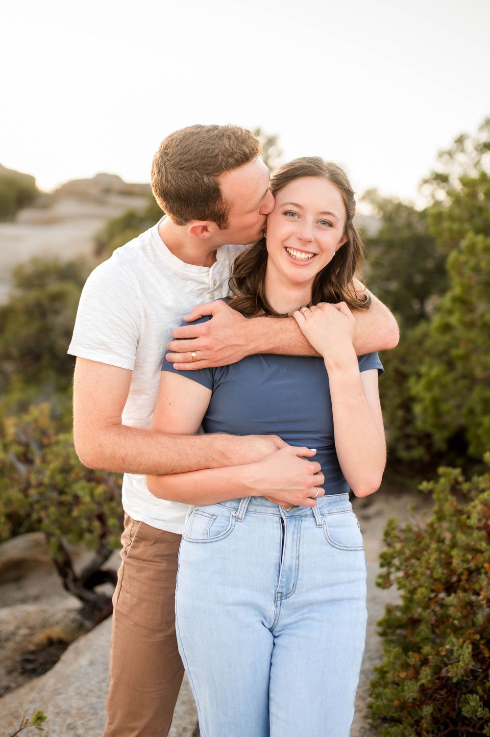 Man kisses smiling woman on cheek while hugging her from behind outdoors.