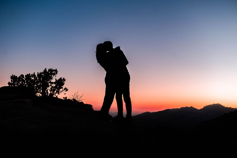 Romantic couple silhouette kissing on a mountain at sunset with colorful orange and blue sky backdrop.