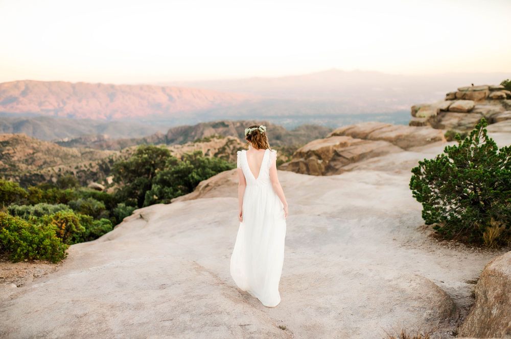 Bride in white gown standing on rocky overlook facing scenic mountain landscape at sunset.