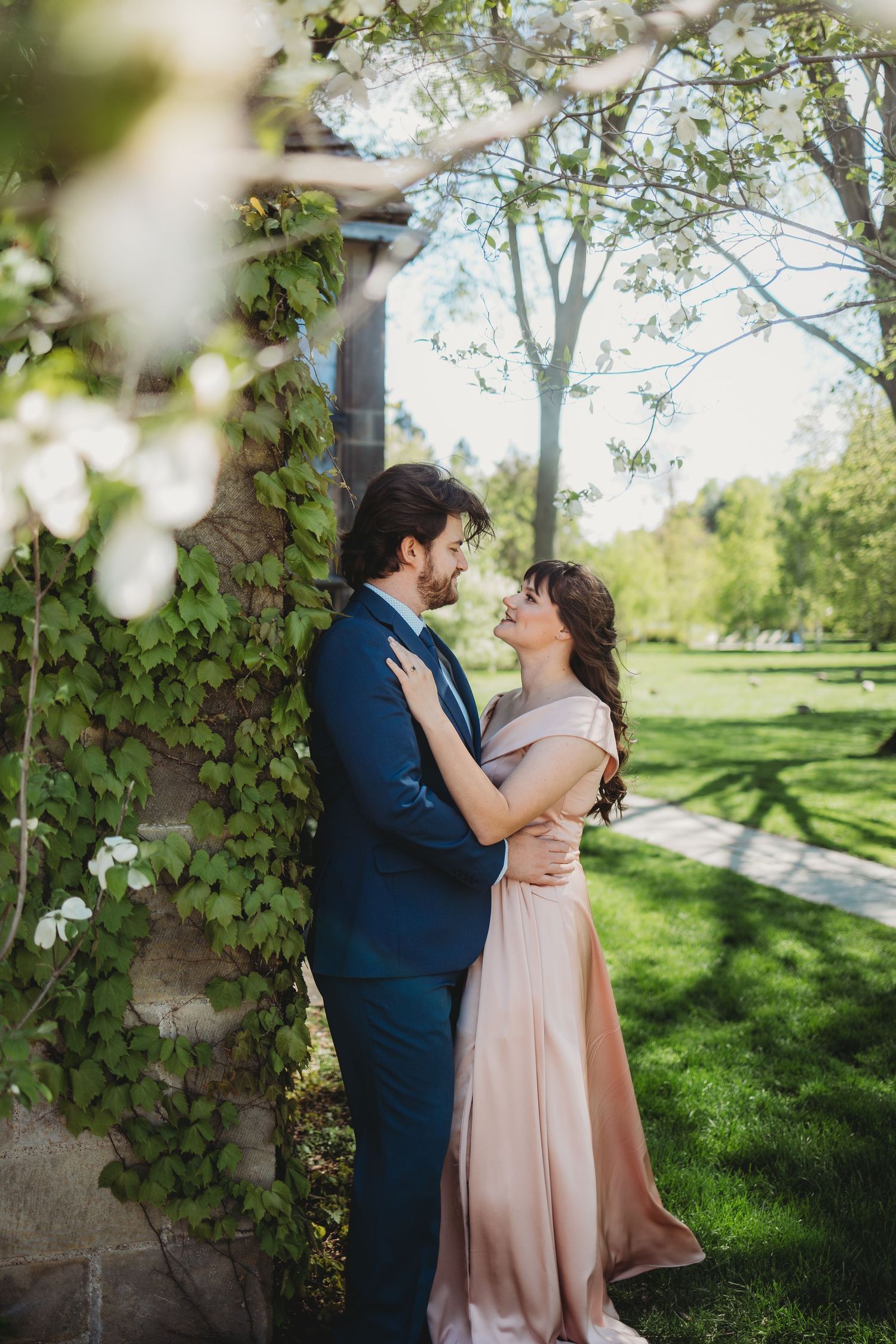 Couple in formal wear embracing in a lush garden with blooming trees and ivy-covered stone wall.