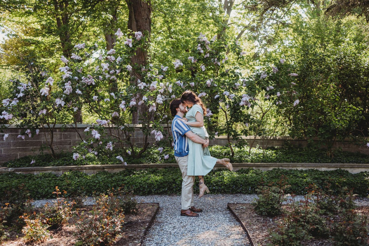 Couple embracing in a lush garden with blooming lilac bushes and green trees in the background.