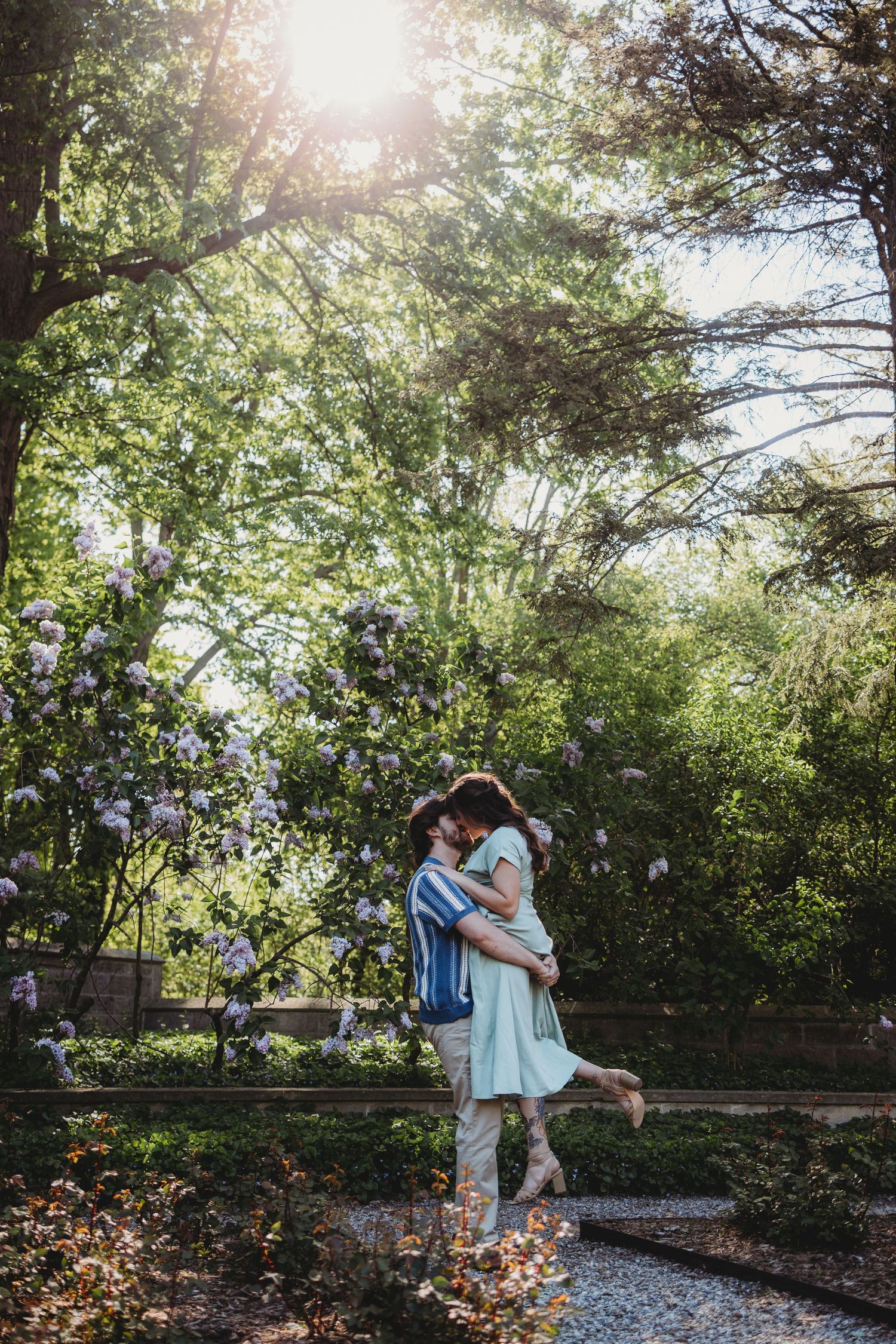 Couple embracing and kissing in a sunlit garden surrounded by blooming lilac bushes and lush green trees.