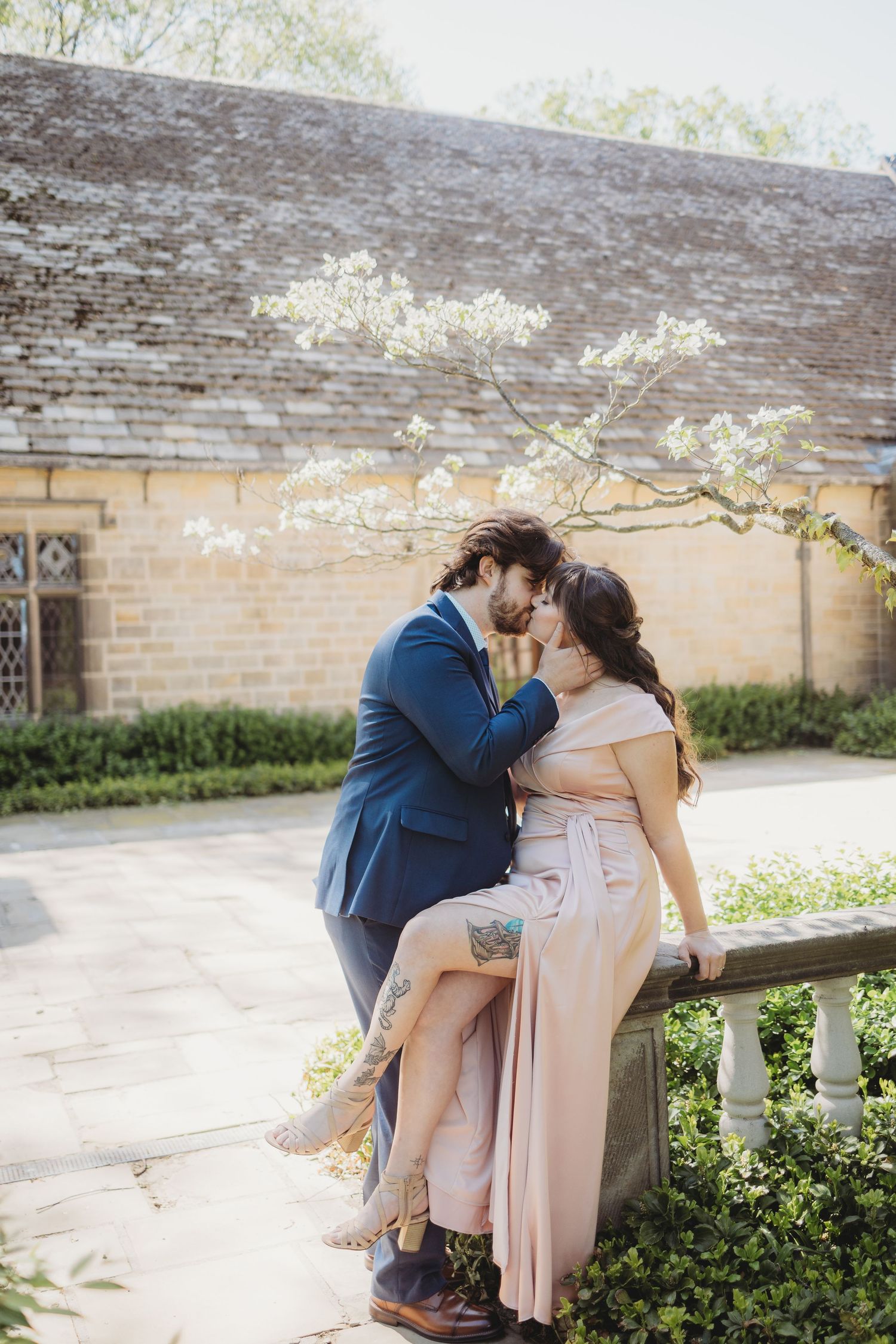 Couple sharing a romantic moment on a stone balustrade in a garden, man in blue suit and woman in blush pink dress.