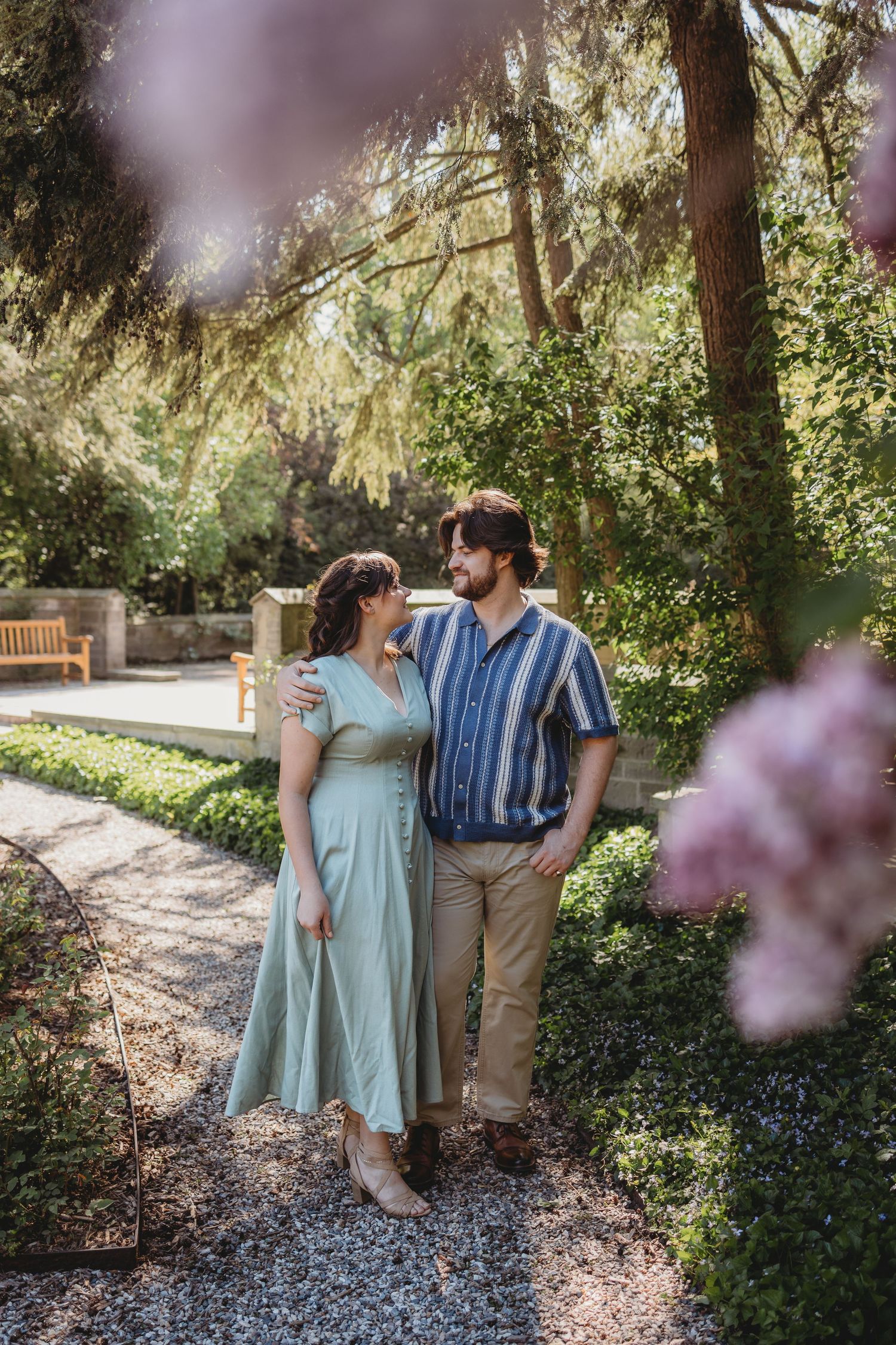 A couple embraces on a garden path surrounded by lush greenery, she in a mint dress, he in a striped shirt.