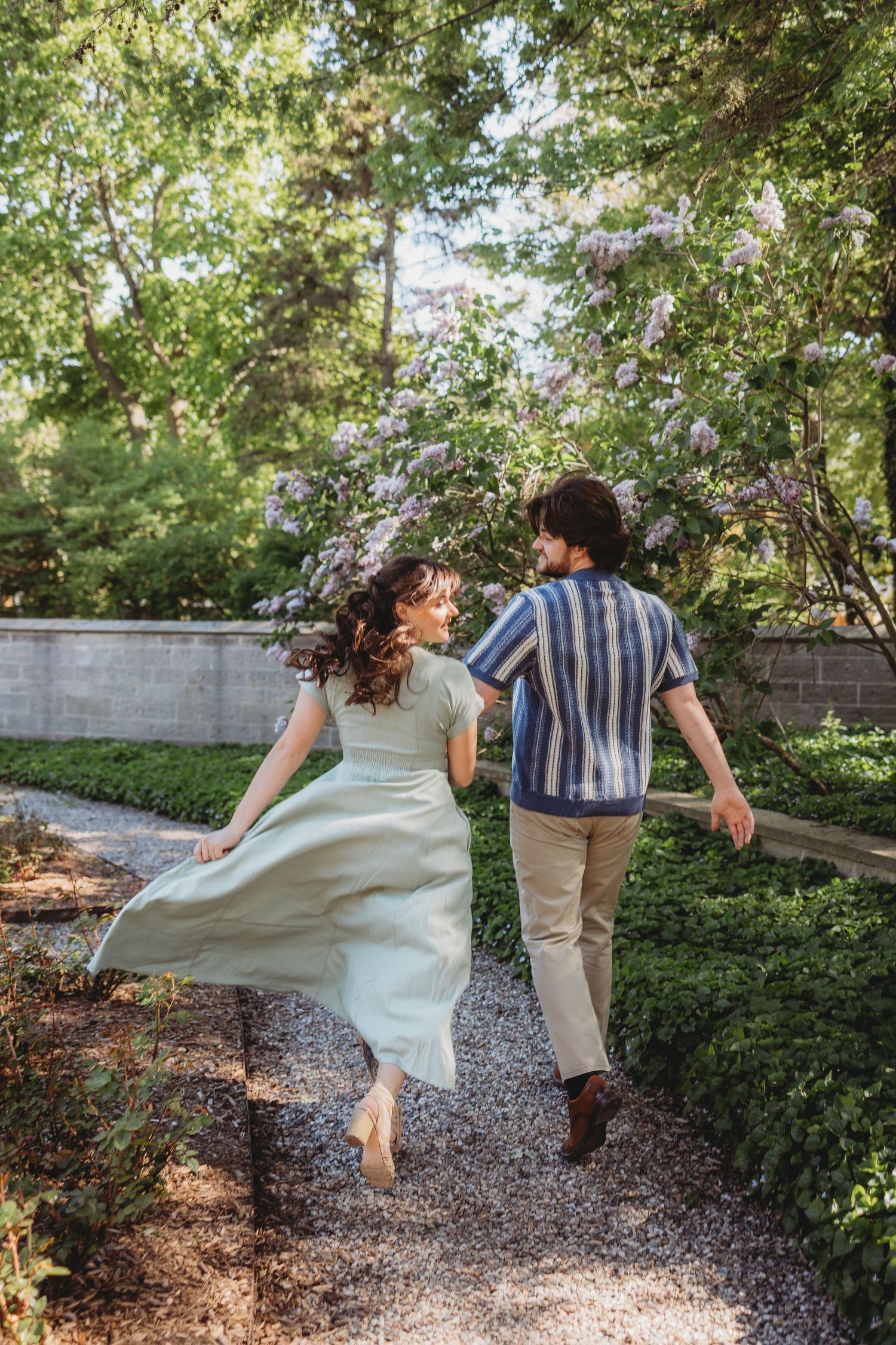 Couple holding hands running through a lush garden path with blooming lilac trees in summer.