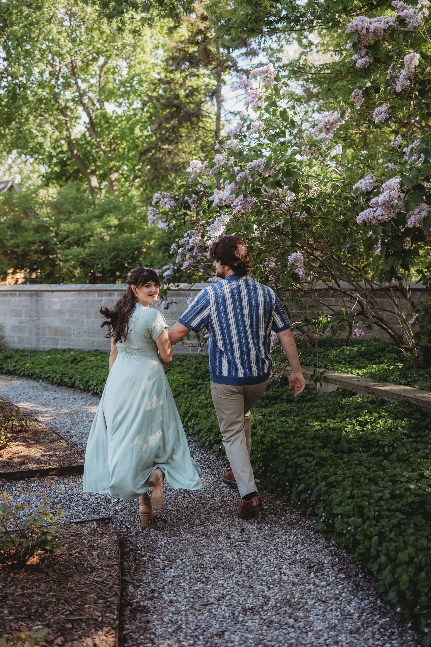 Couple holding hands walking on a gravel garden path surrounded by lush greenery and blooming lilac bushes.
