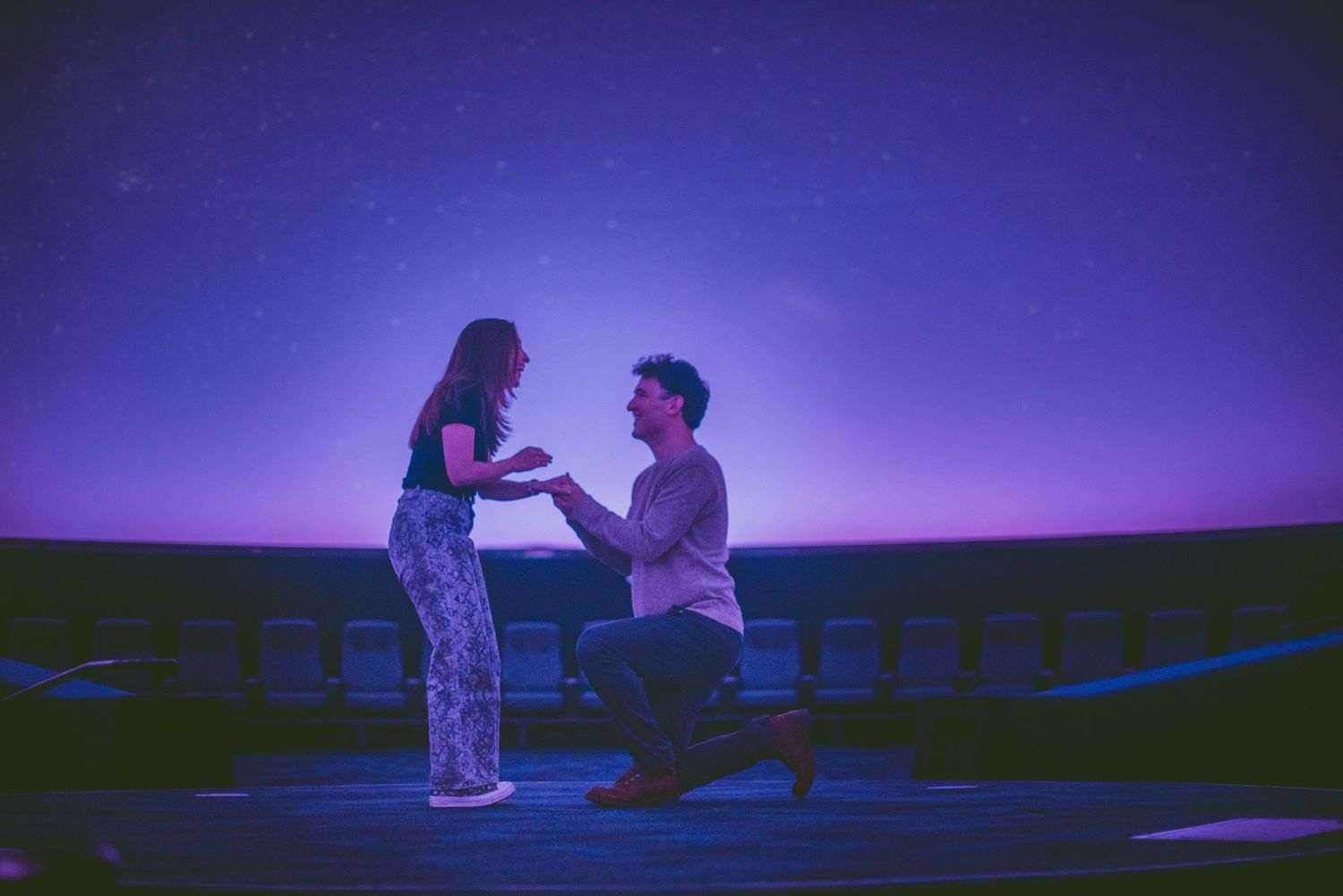 Romantic marriage proposal under a starry purple sky, man kneeling before woman outdoors at night.