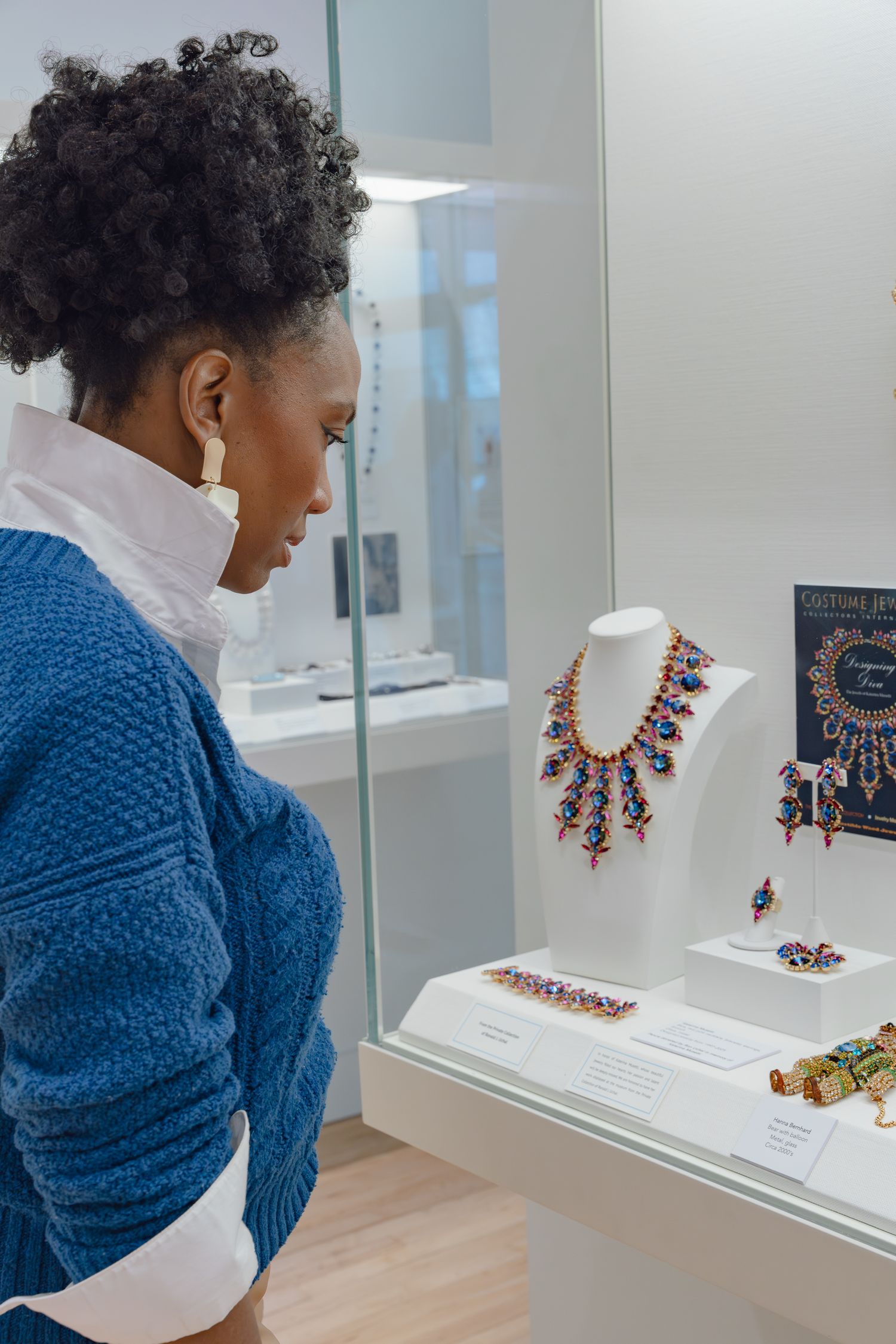 A woman in a blue sweater admires colorful gemstone jewelry on display in a museum glass case.