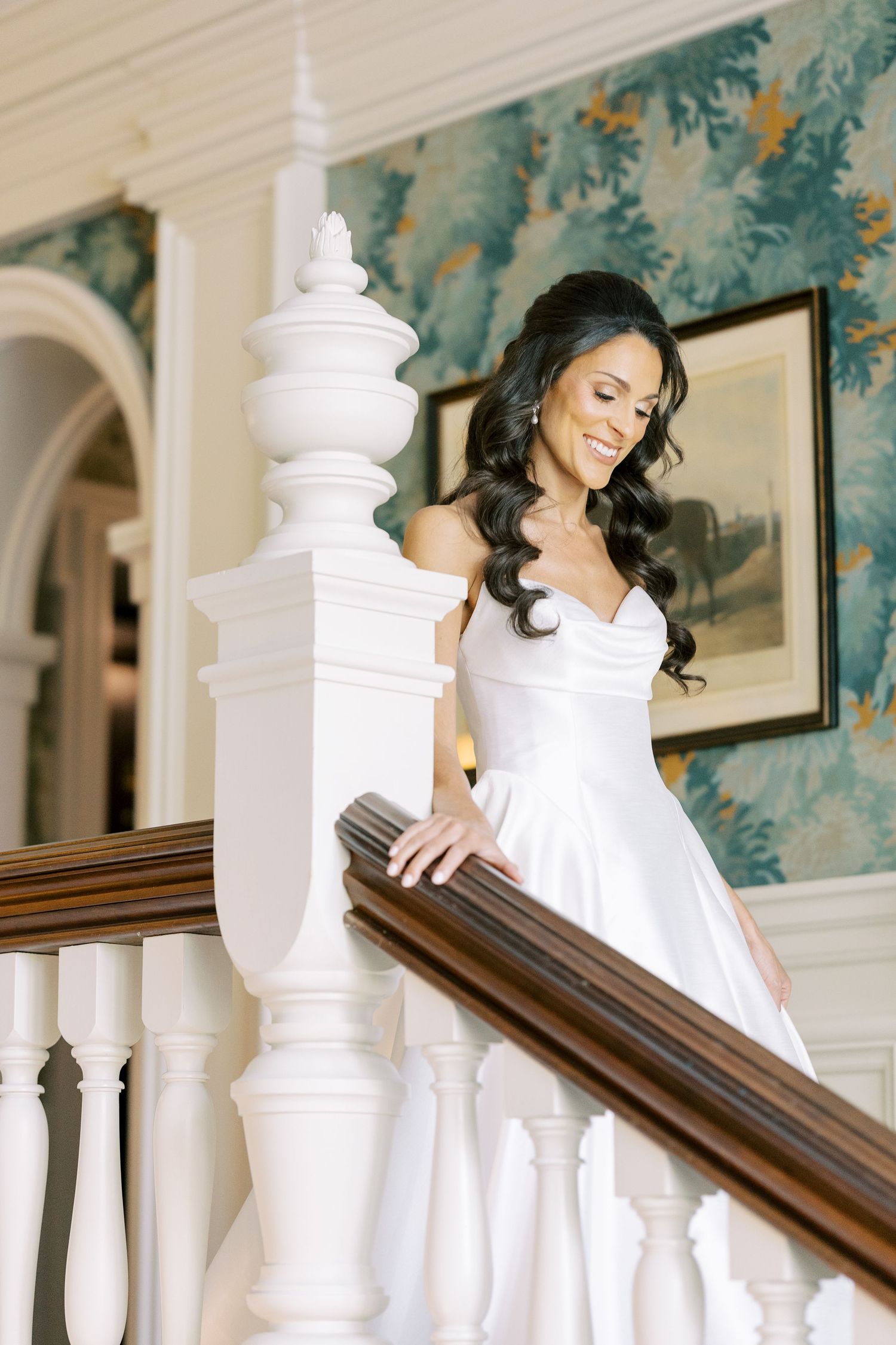 Smiling bride in white wedding gown descending elegant staircase with white balusters and floral wallpaper backdrop.