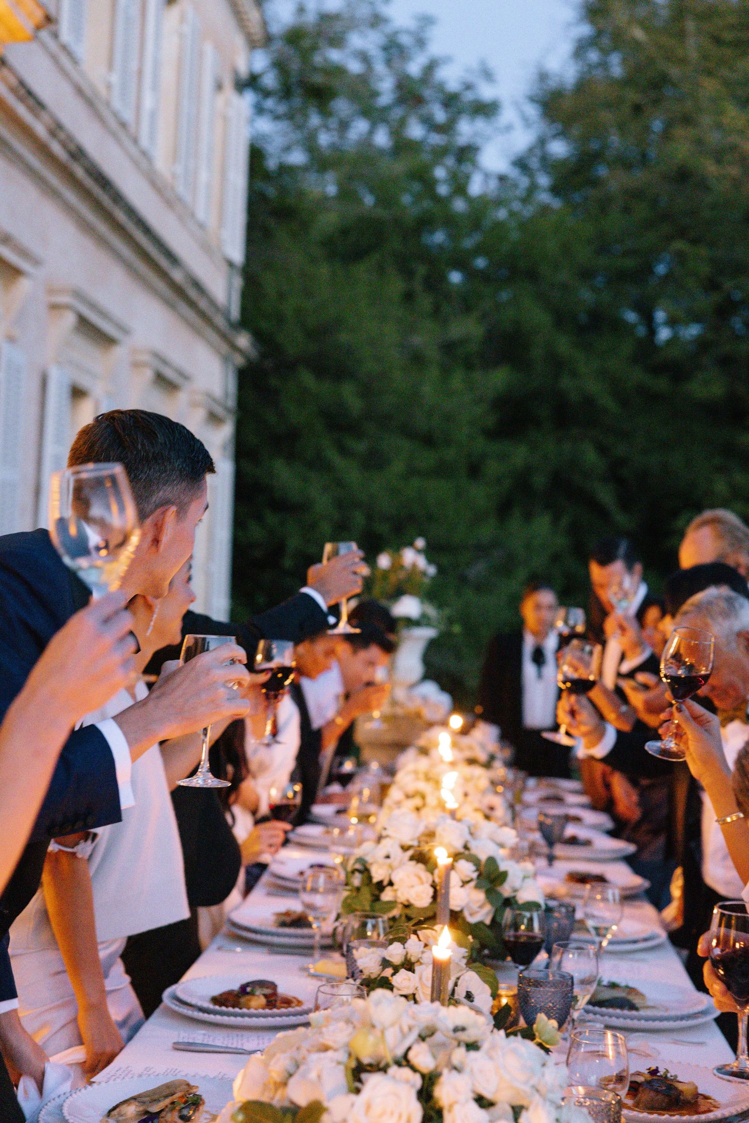 Elegant outdoor wedding dinner toast with guests raising wine glasses along a candlelit table adorned with white floral centerpieces.