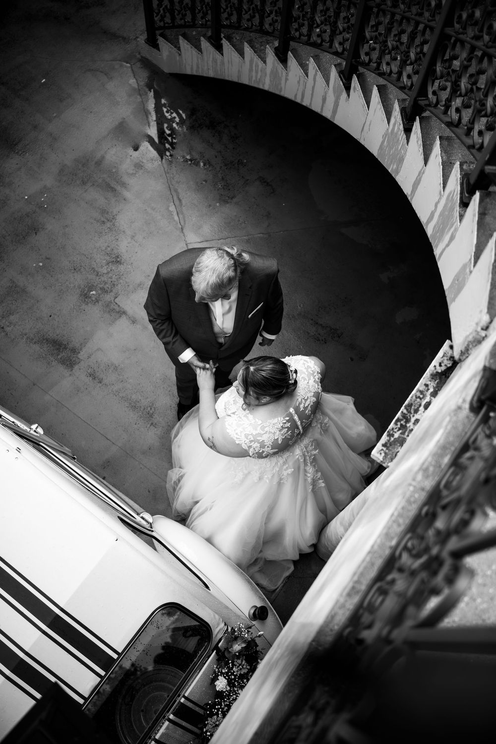 Aerial black and white view of bride and groom near vintage car under spiral staircase arch.