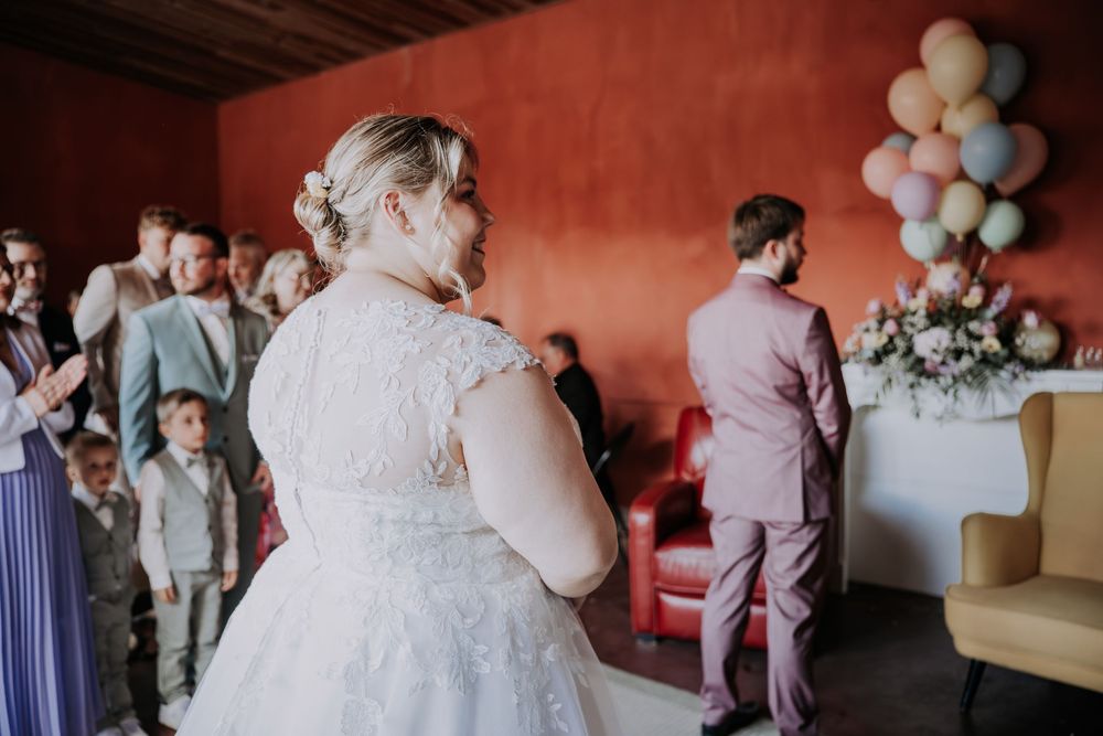 Bride in lace wedding gown walks toward groom at indoor ceremony with pastel balloons.