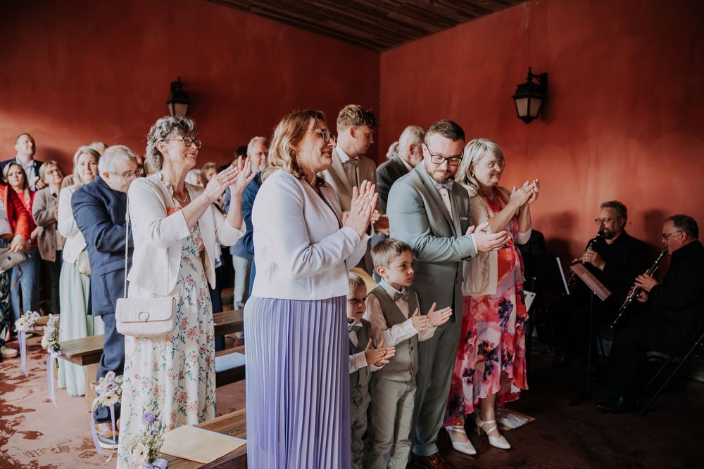 Wedding guests clapping and raising hands in celebration inside a red-walled venue during a ceremony.