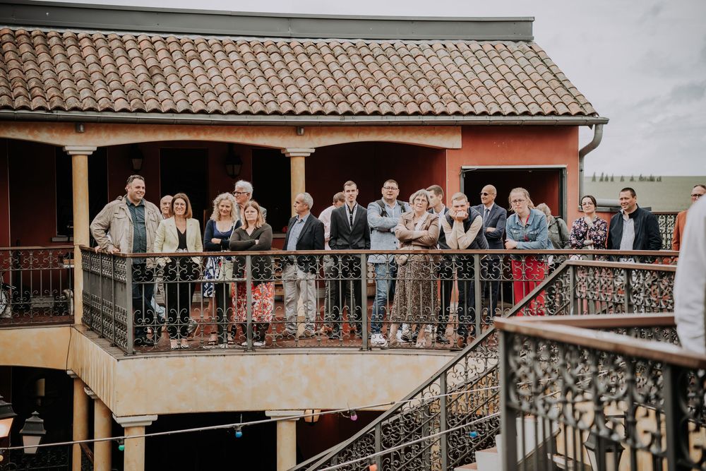 Group of wedding guests gathered on a rustic balcony of a Mediterranean-style venue with ornate iron railings.