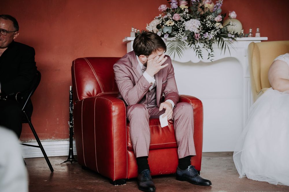 Emotional groom in pink suit sitting in red armchair, covering face with hands during wedding ceremony.