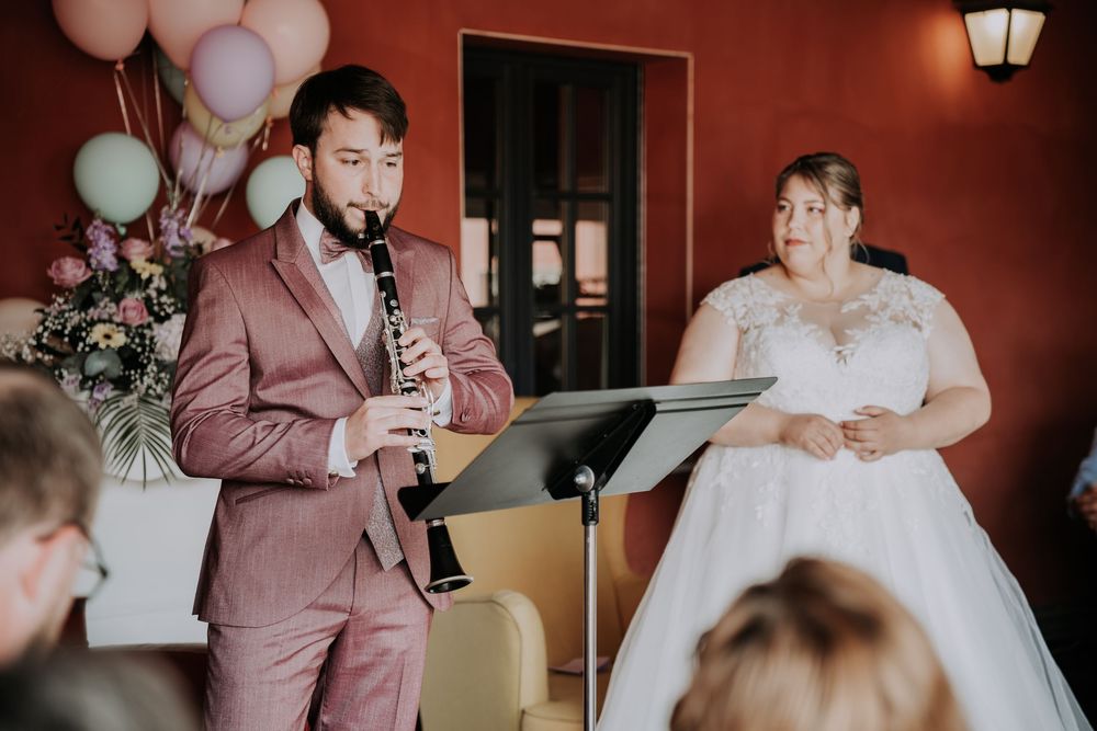 Groom plays clarinet beside bride in wedding gown at indoor ceremony with pastel balloons.
