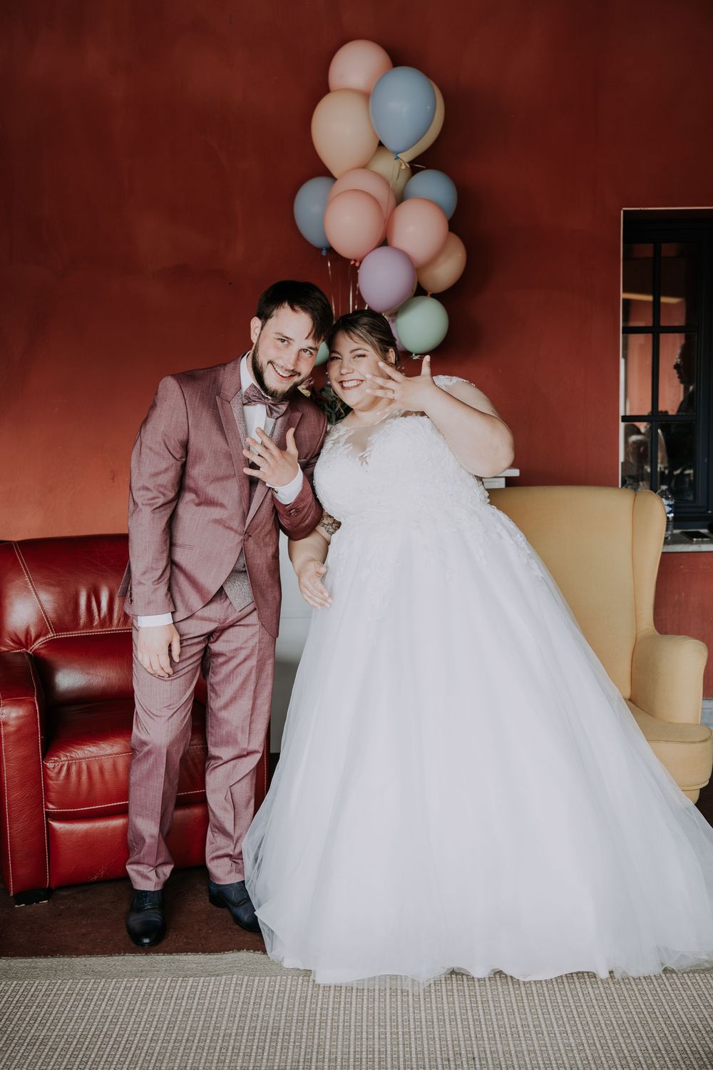 Joyful newlyweds pose with colorful balloons, groom in pink suit and bride in white ball gown, against a red wall.