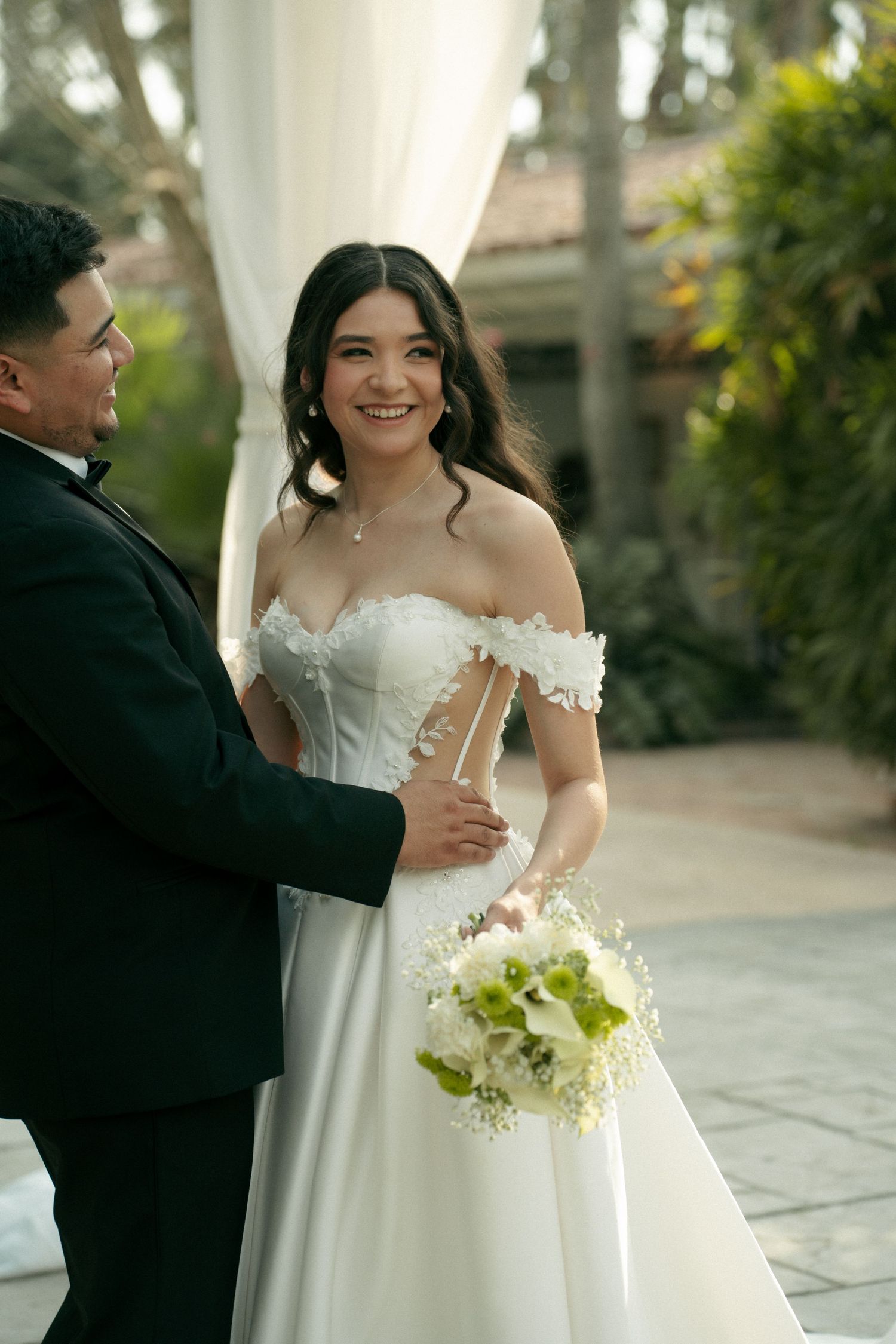 Smiling bride in off-shoulder white gown holds bouquet beside groom in tuxedo at outdoor wedding ceremony.