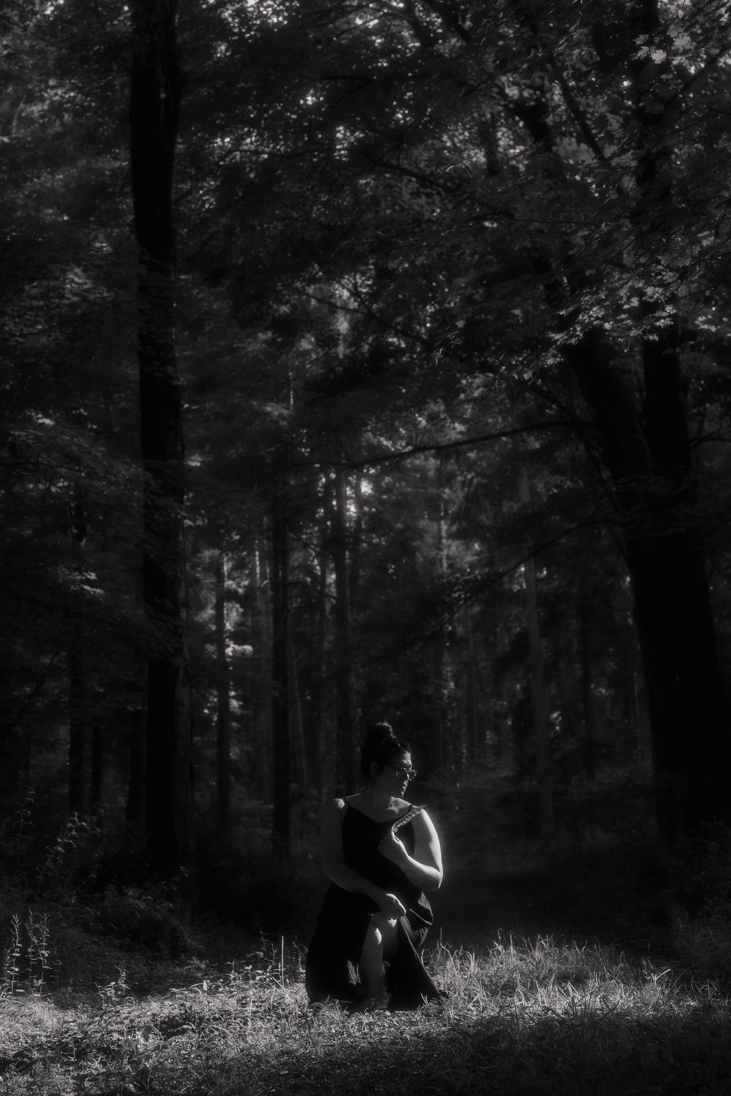 Black and white photo of a woman in a dark dress standing in a moody, sunlit forest clearing.