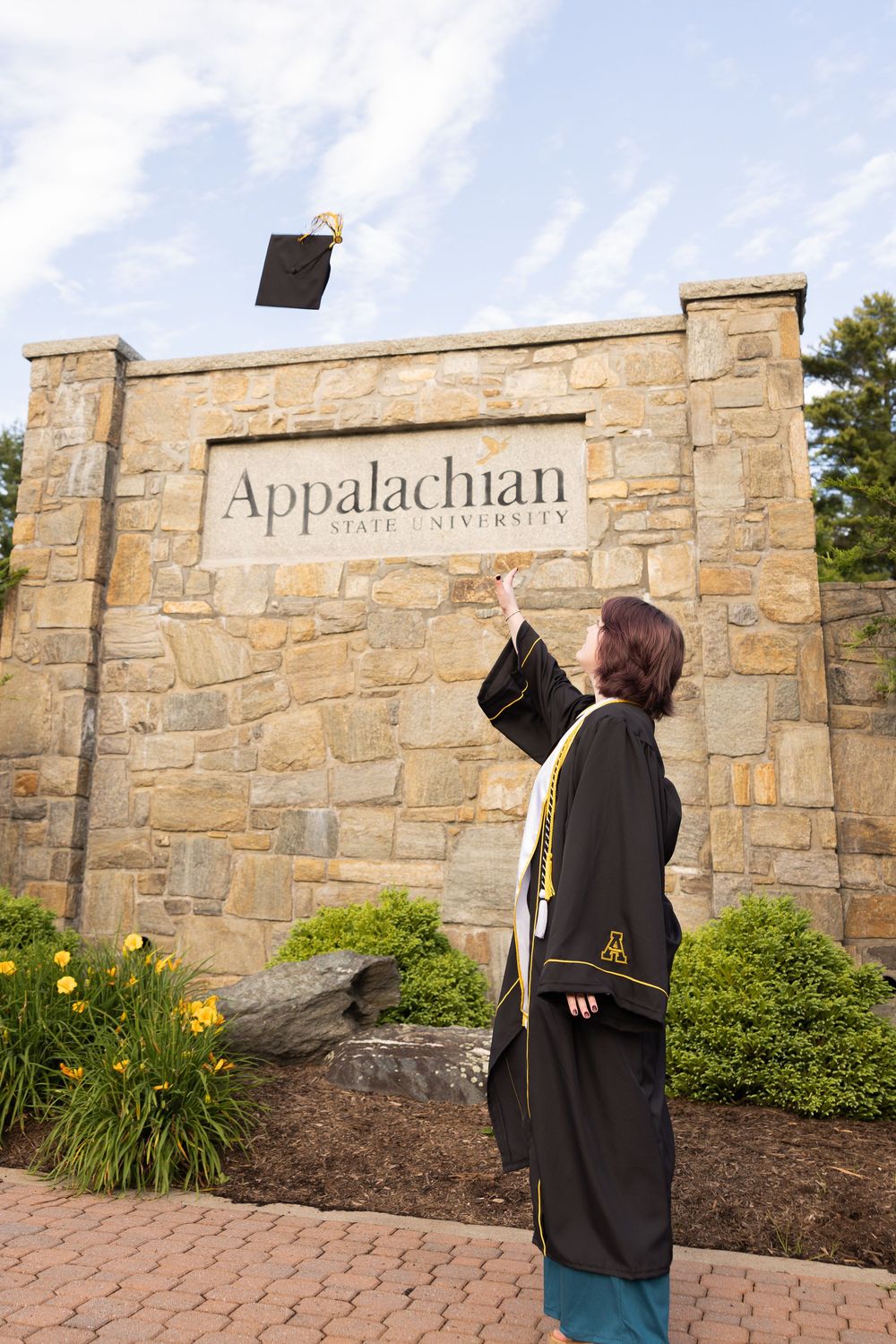 Graduate in cap and gown tossing mortarboard in front of Appalachian State University stone entrance sign.