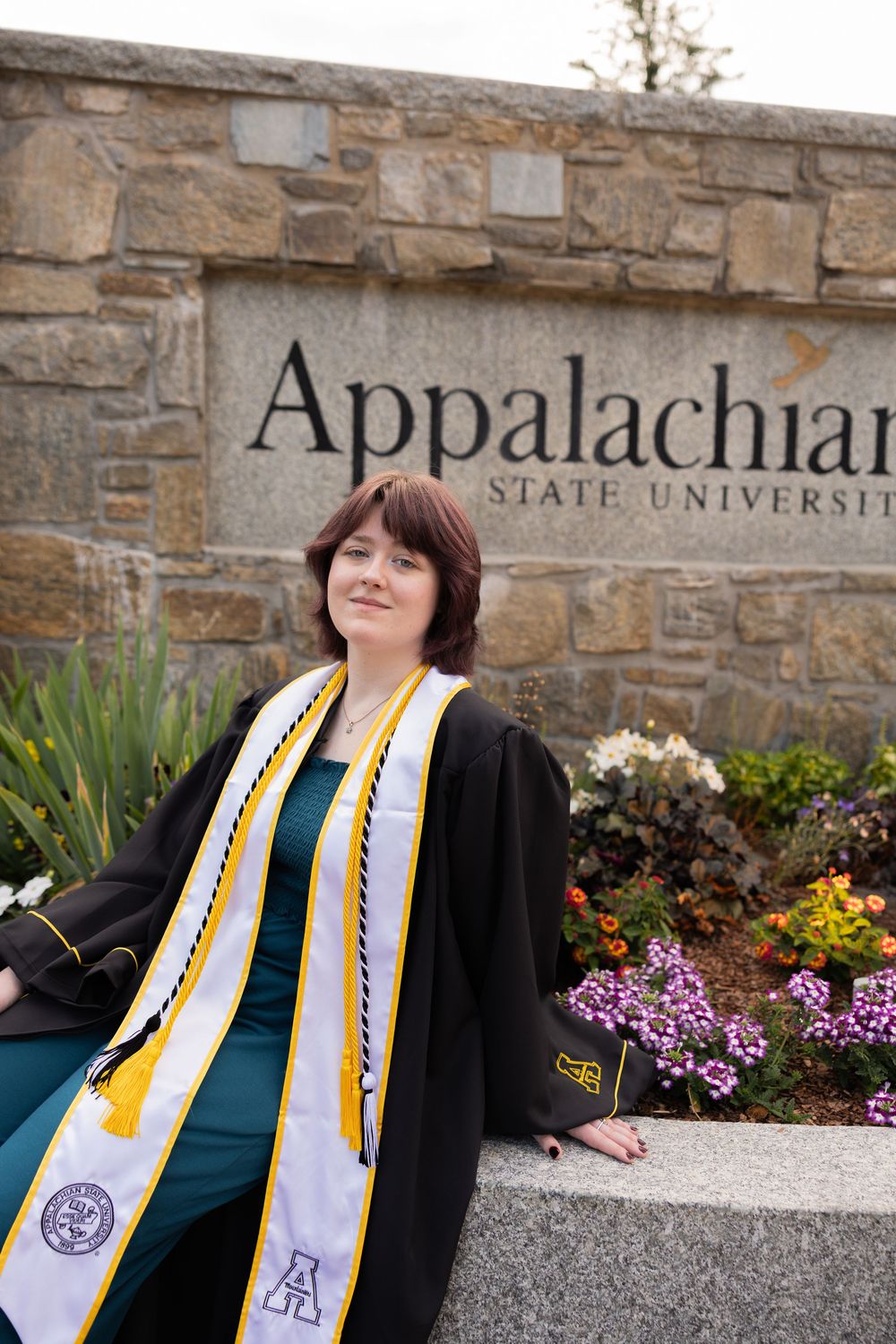 Female graduate in cap and gown poses by Appalachian State University entrance sign with flowers.