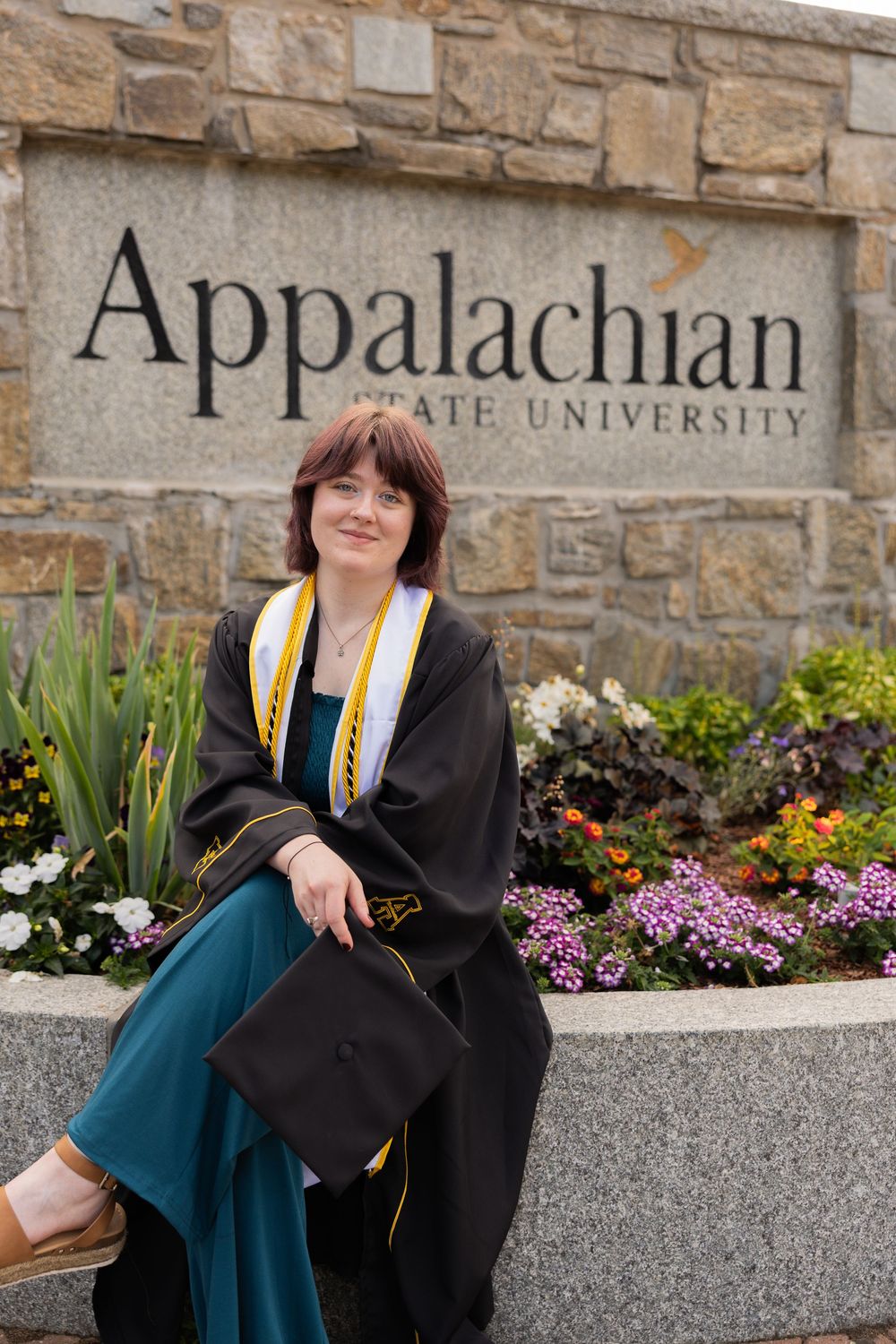 Graduate in cap and gown sits before Appalachian State University sign.