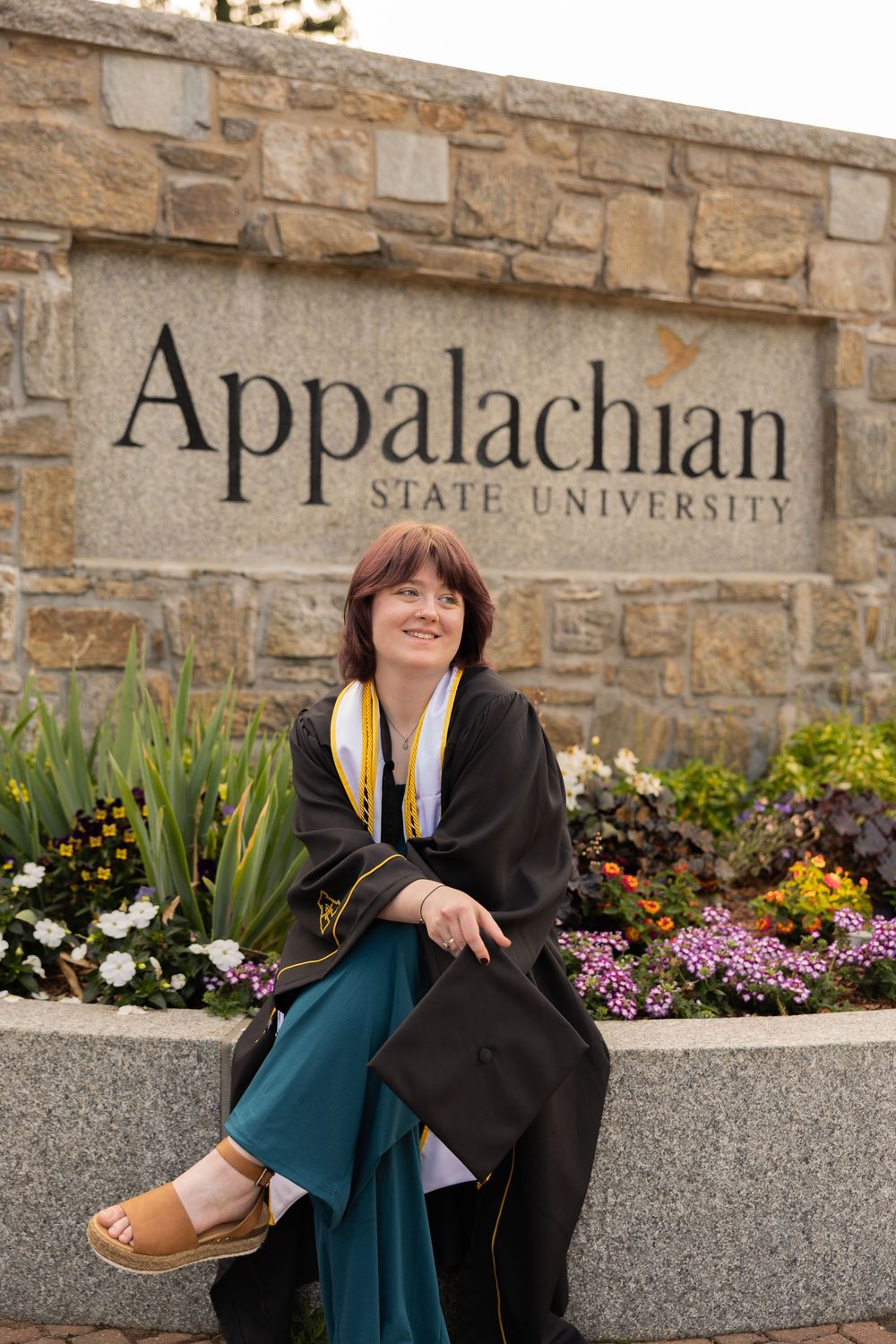 A smiling graduate in black cap and gown sits in front of Appalachian State University stone sign surrounded by colorful flowers.