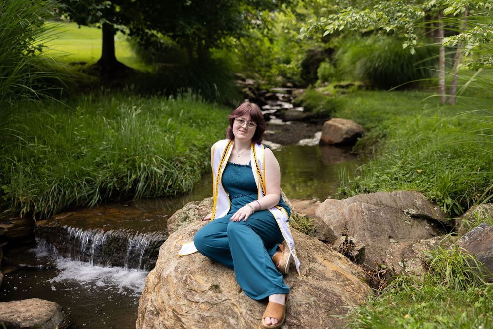 Woman in teal jumpsuit sitting on rock by a peaceful stream surrounded by lush green trees and grass.