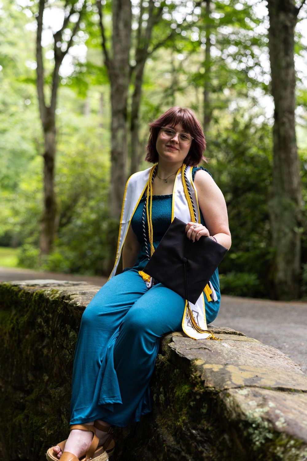 A woman in a teal dress with graduation regalia sits on a stone wall surrounded by lush green trees.