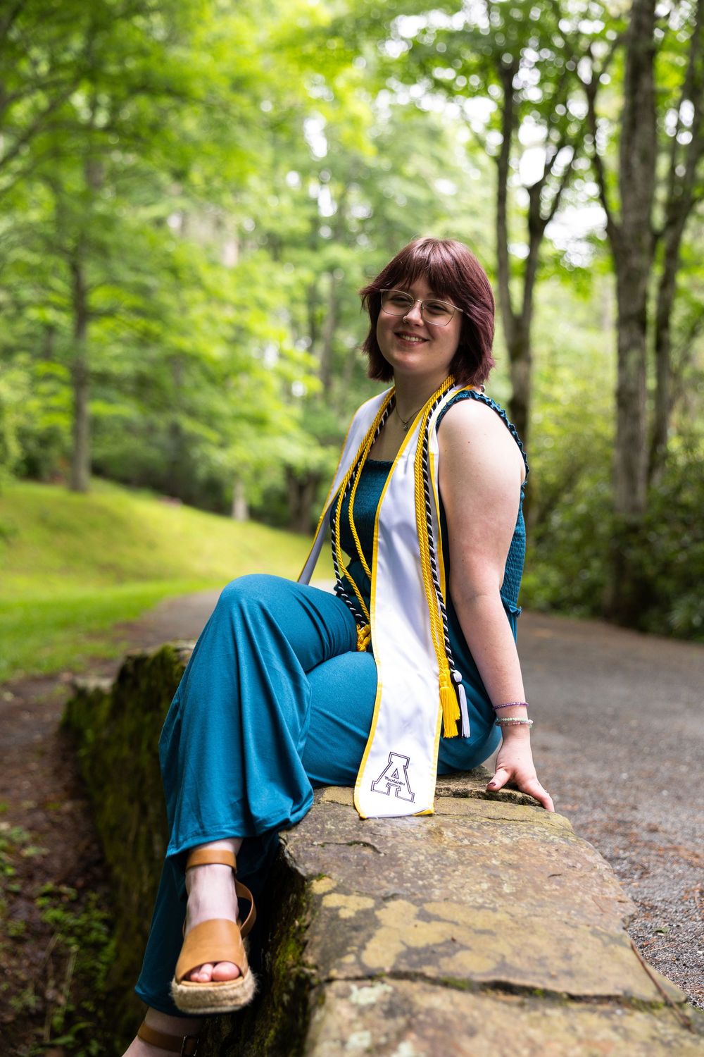 Young woman in teal jumpsuit and graduation stole sits on stone wall in lush green forest park setting.