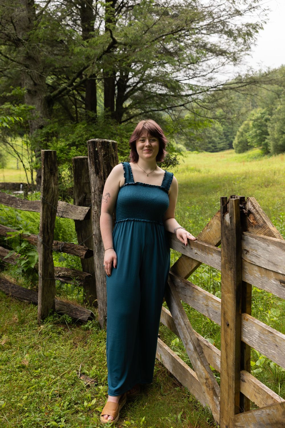 Woman in teal jumpsuit posing by rustic wooden fence in lush green countryside setting.