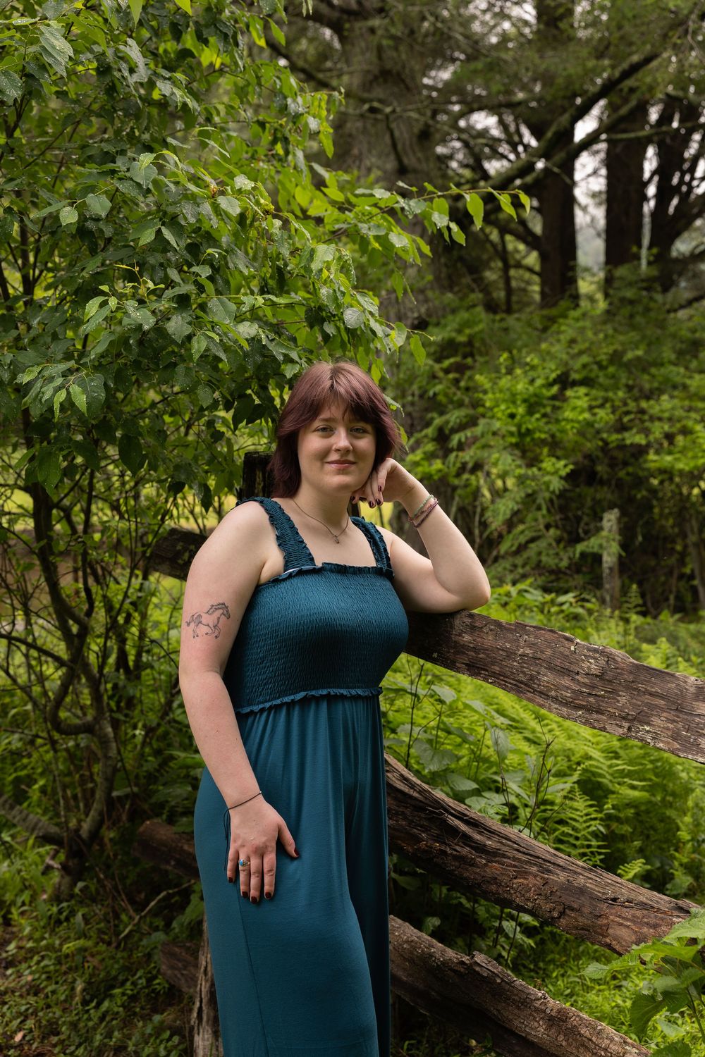 Woman in teal maxi dress poses by rustic wooden fence surrounded by lush green forest foliage.