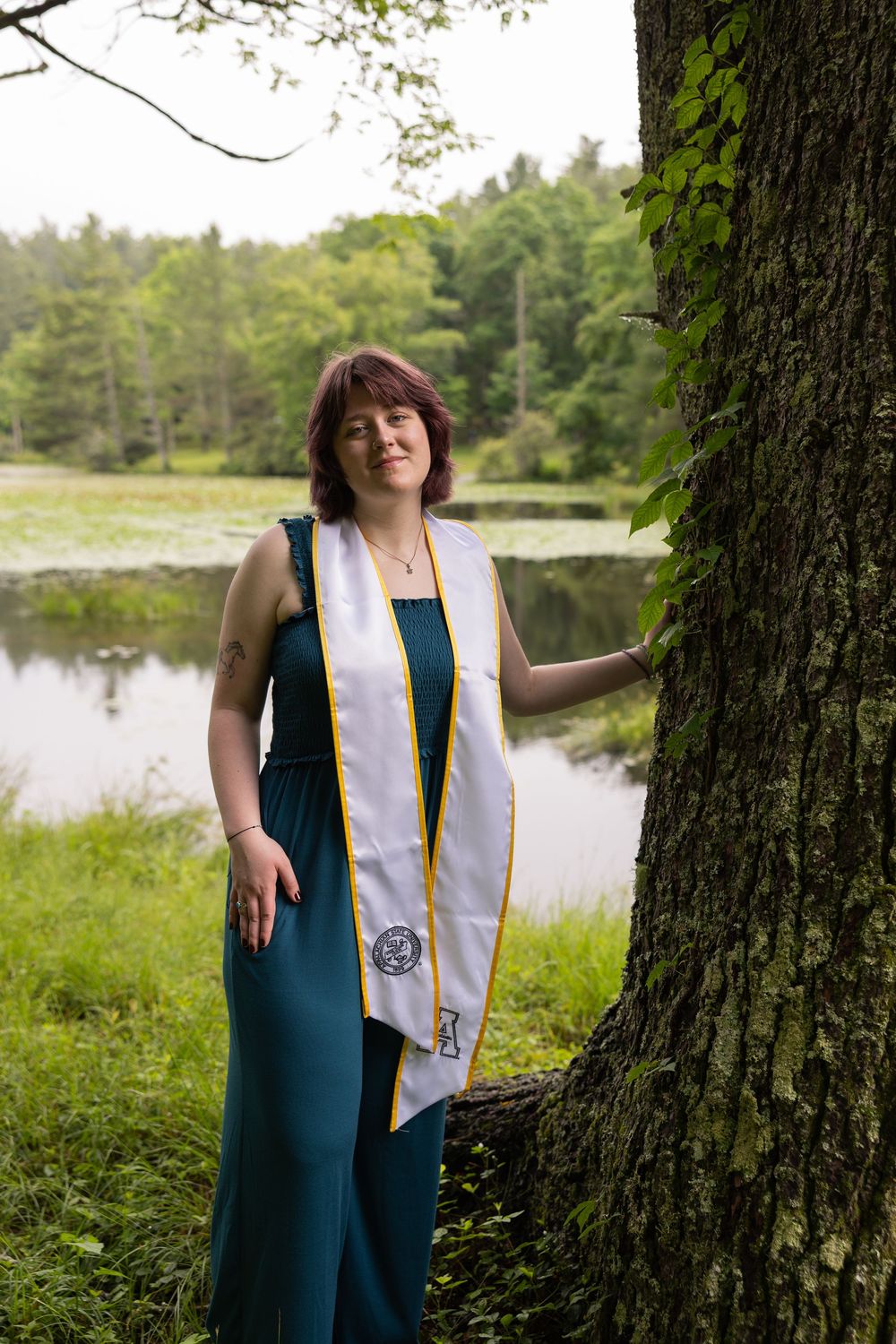 Woman in teal dress and graduation stole poses by tree near pond.