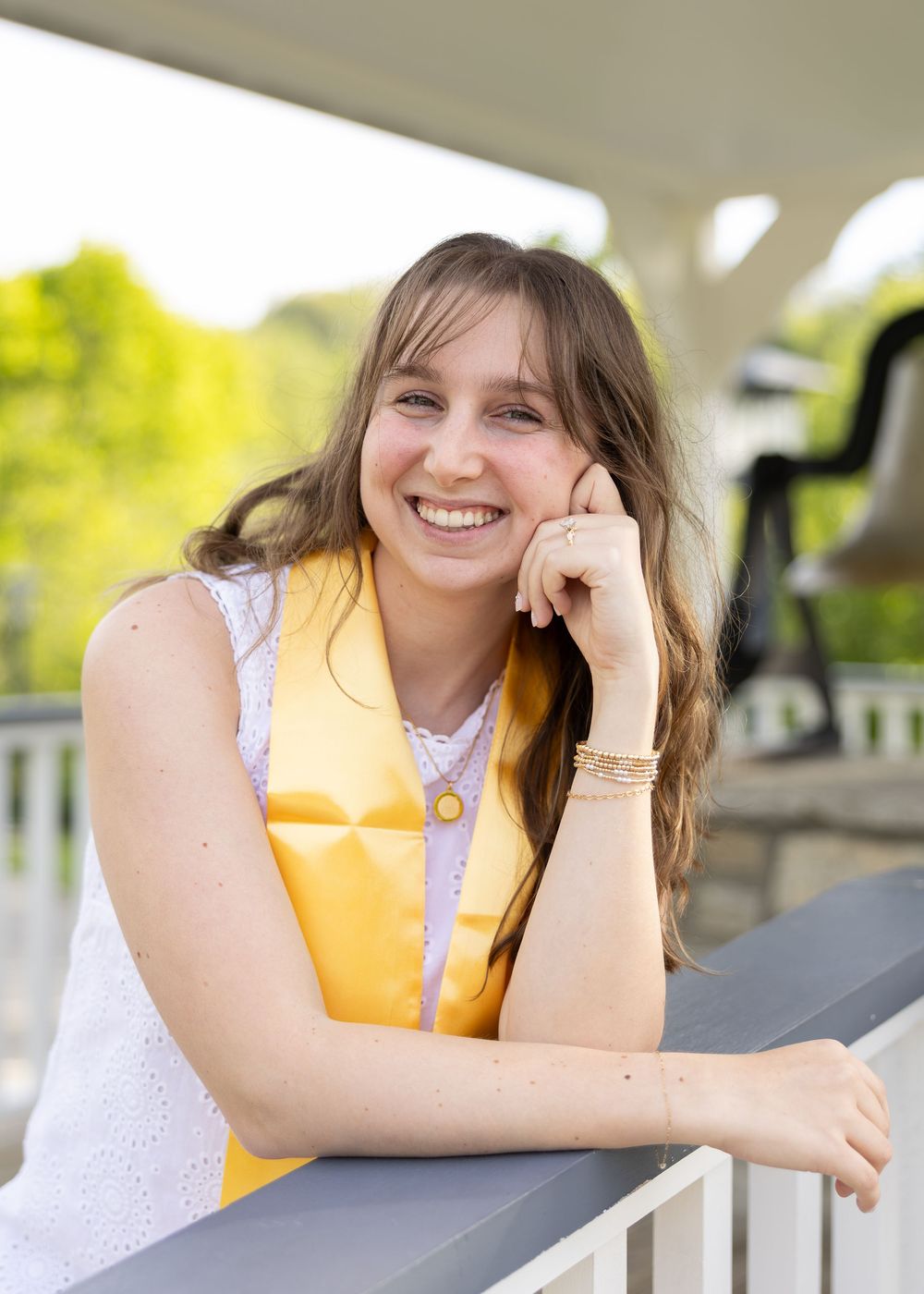 Smiling young woman in graduation regalia leaning on a railing outdoors during a sunny day.