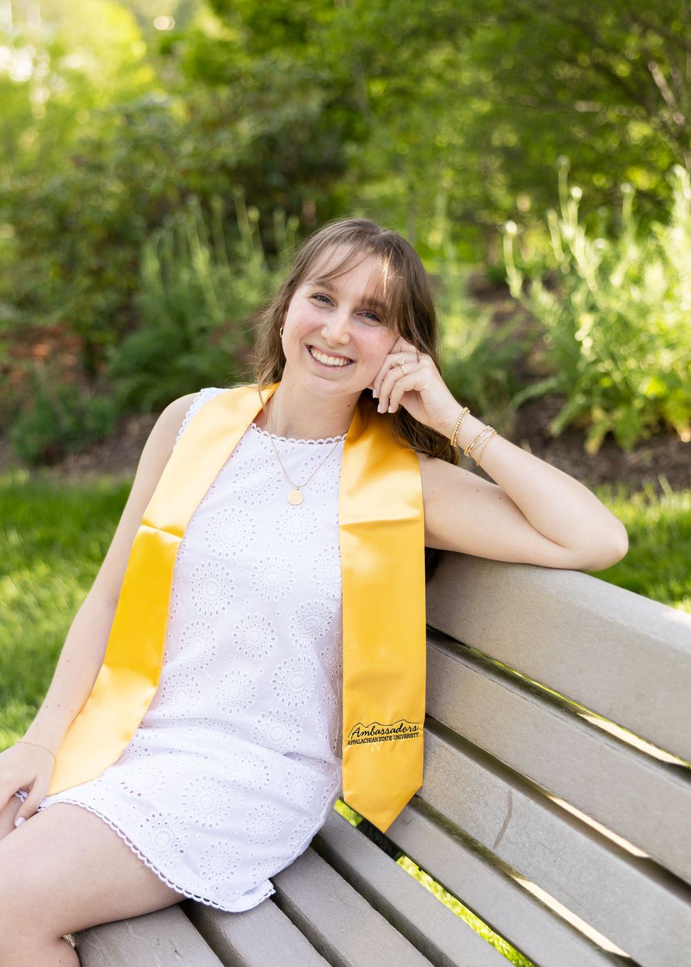 Smiling young woman in white dress and gold graduation stole sitting on a bench outdoors among green trees.