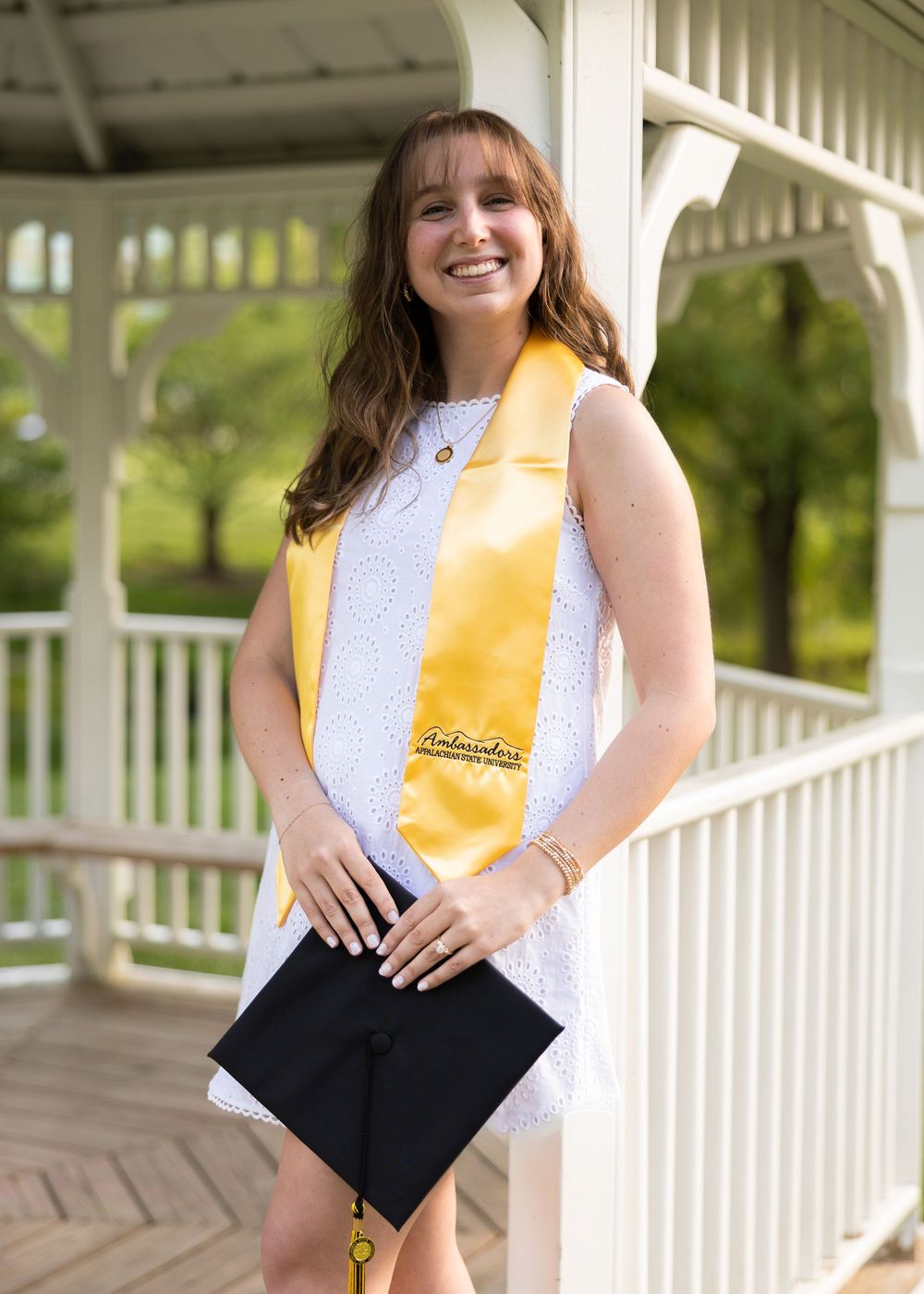 Smiling female graduate in white dress with gold honor stole holding cap at outdoor gazebo.