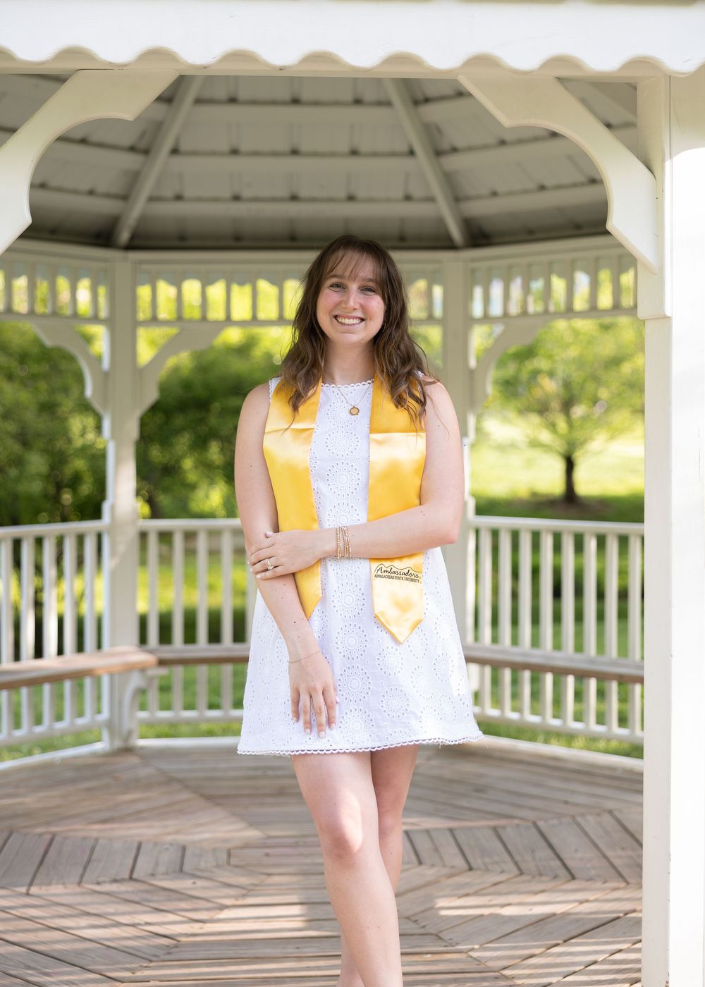 Happy female graduate in white lace dress and gold stole posing inside white wooden gazebo.