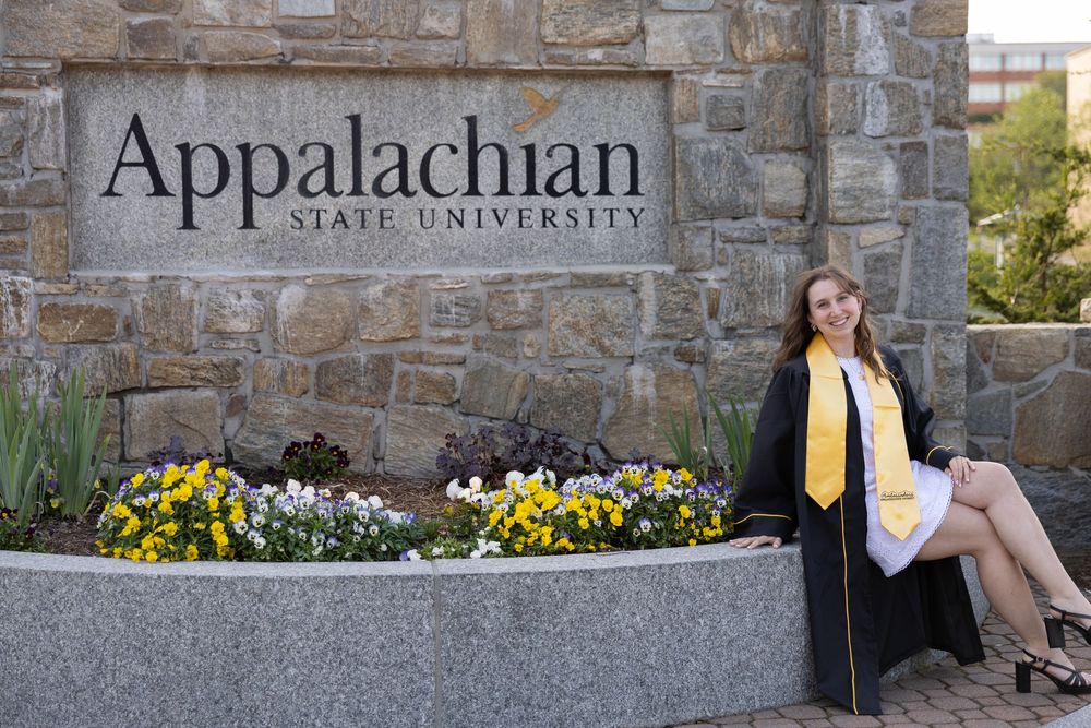 Graduate in cap and gown poses by Appalachian State University stone sign surrounded by colorful flowers.