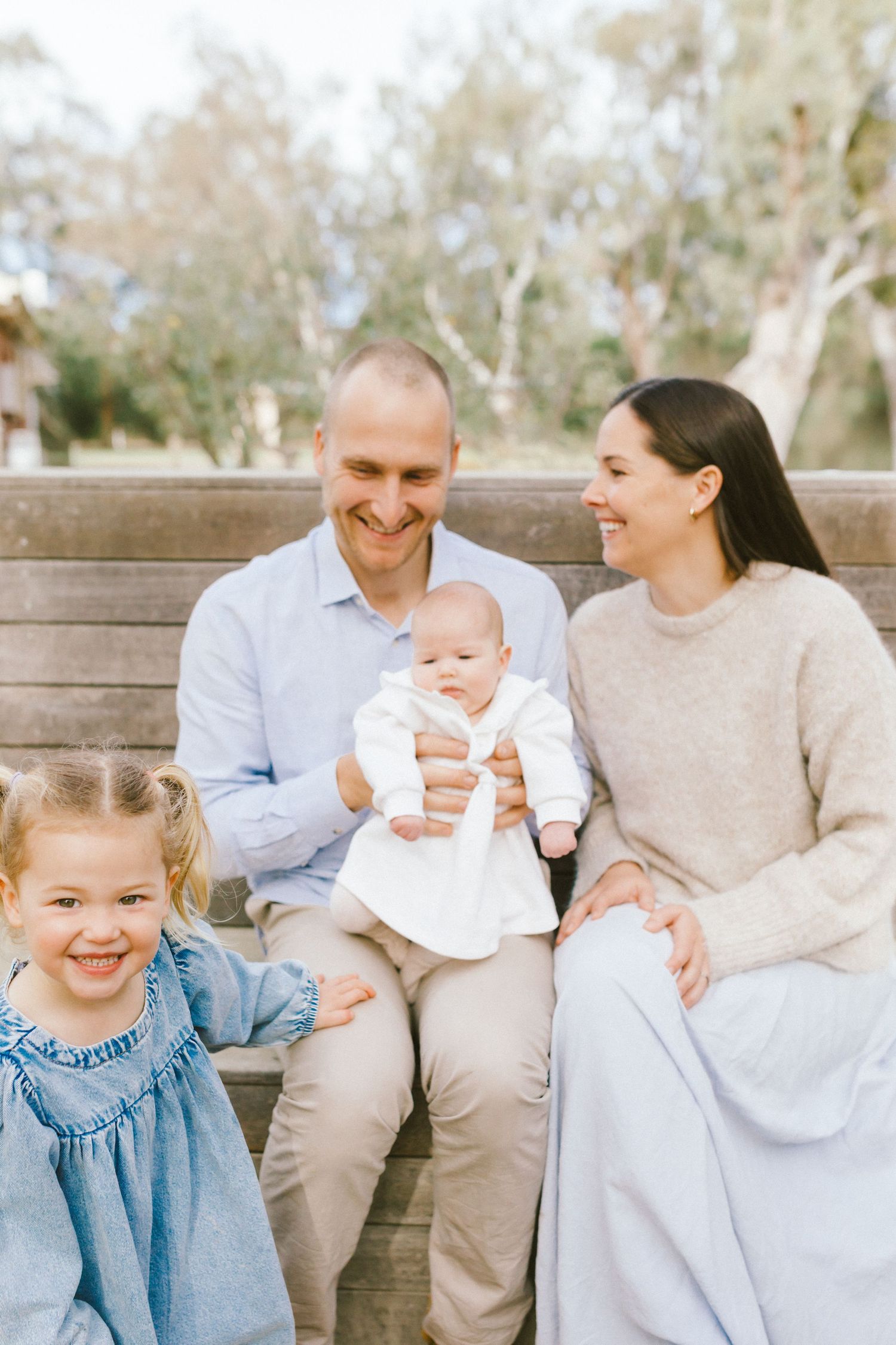 Happy family of four sitting on a wooden bench outdoors, parents smiling with toddler girl and baby in white outfit.