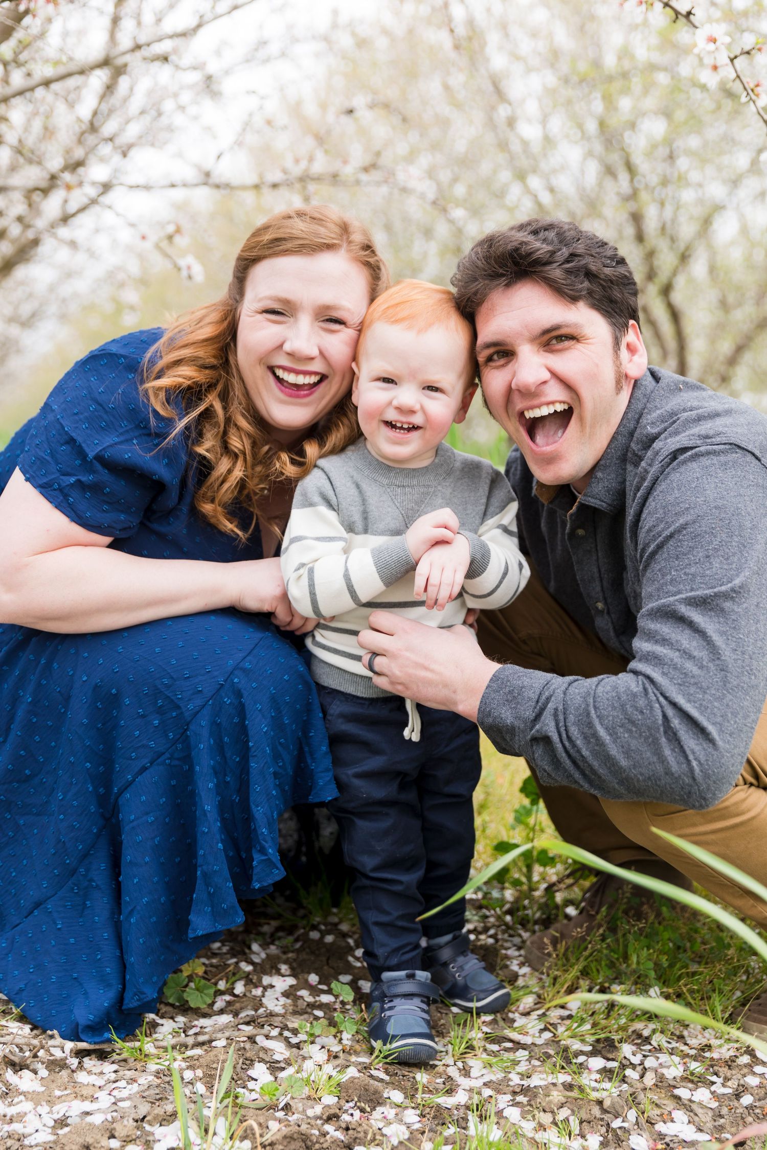 Happy family of three posing outdoors in spring orchard, parents laughing with red-haired toddler boy among white blossoms.