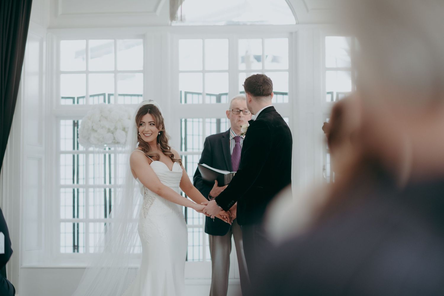 Bride and groom exchange vows at indoor wedding ceremony with officiant, large windows in elegant white venue.