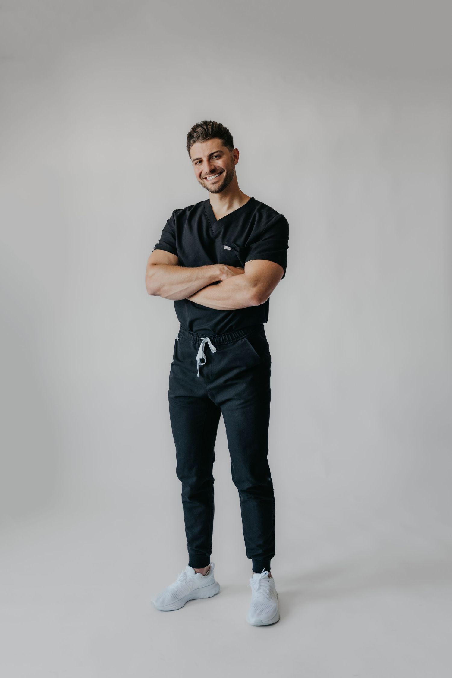 Smiling man in black medical scrubs with arms crossed, wearing white sneakers, posing against a gray background.